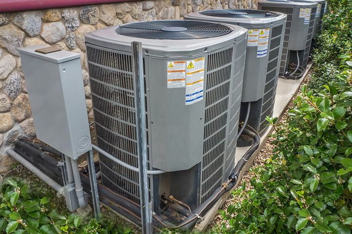 Several outdoor air conditioning units lined up against a stone wall, with electrical box and bushes.