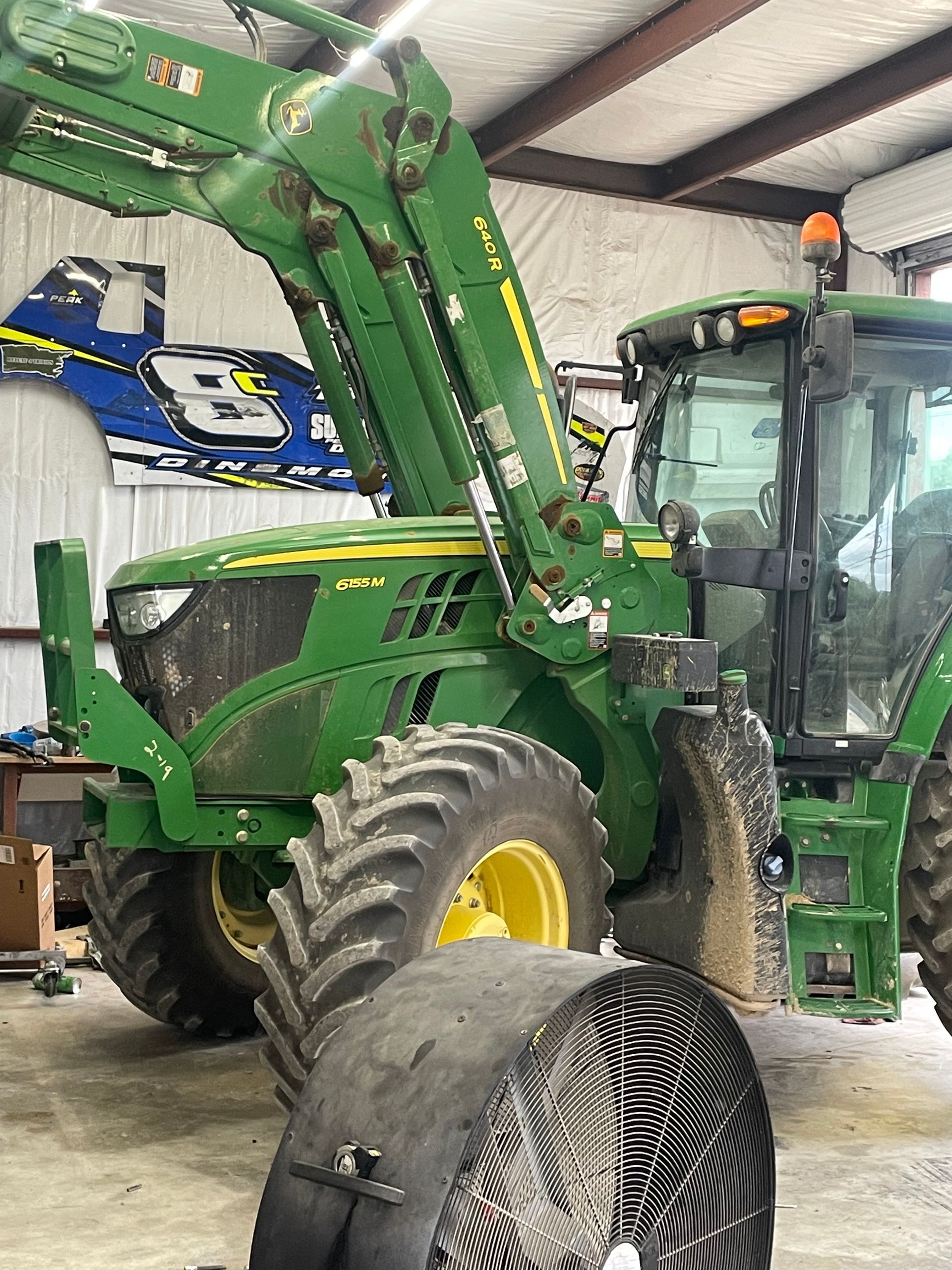 A green tractor is parked in a garage next to a fan.