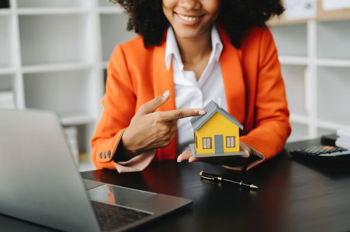 Person in orange blazer pointing at a small yellow house model beside a laptop
