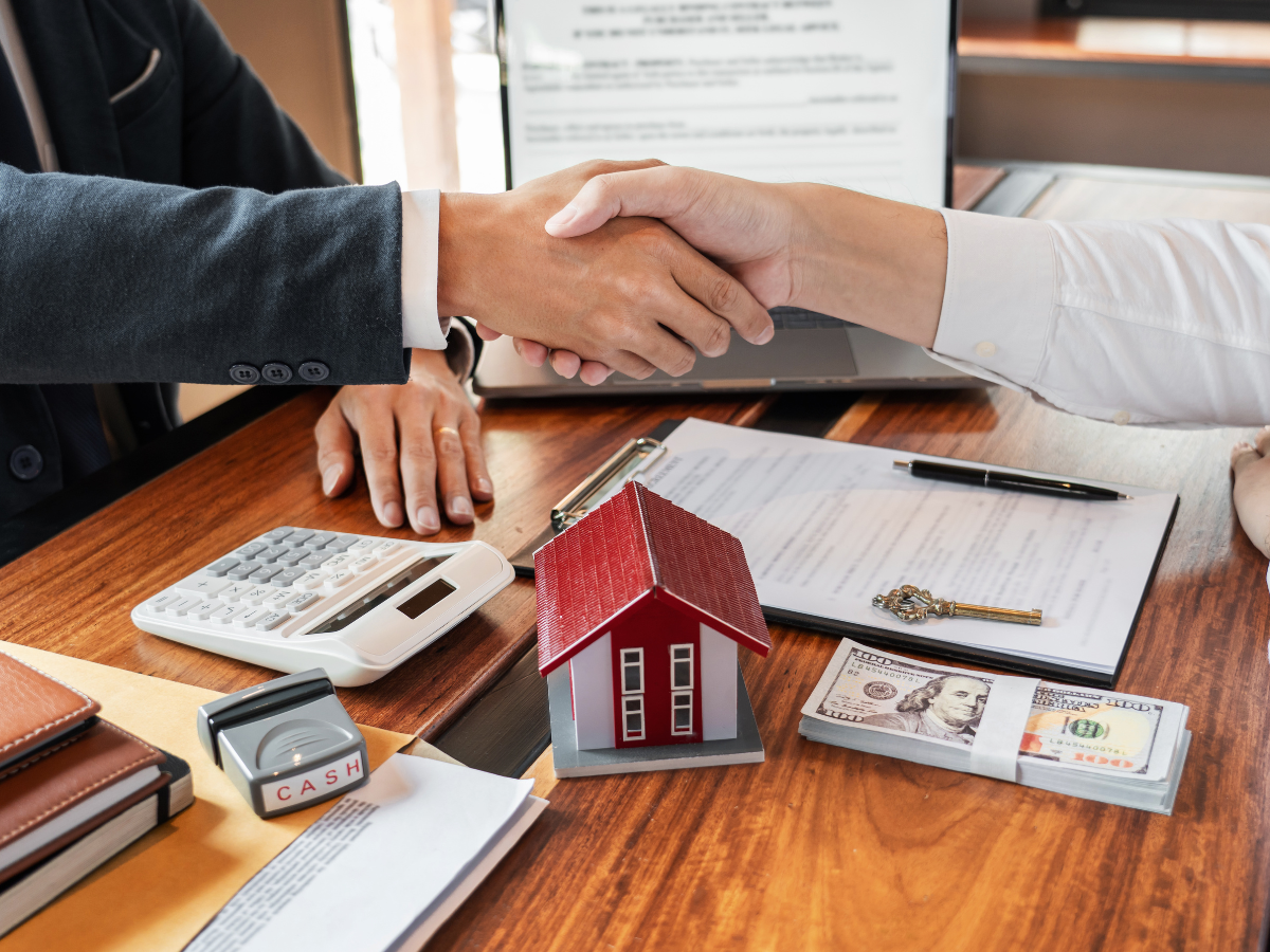 Two people shaking hands over a table with a house model, documents, and money.