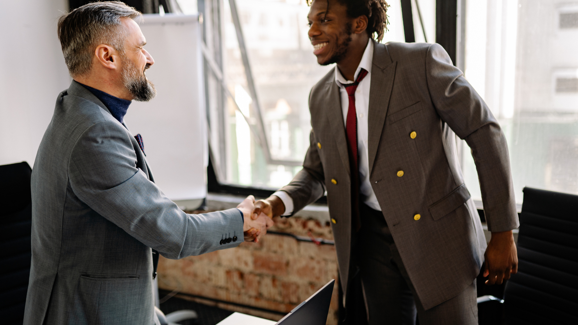 Two men in suits shaking hands in an office, smiling.