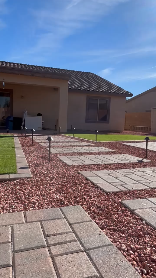 A backyard pathway of stepping stones and red gravel leads to a beige house, lit by pathway lights under a blue sky.