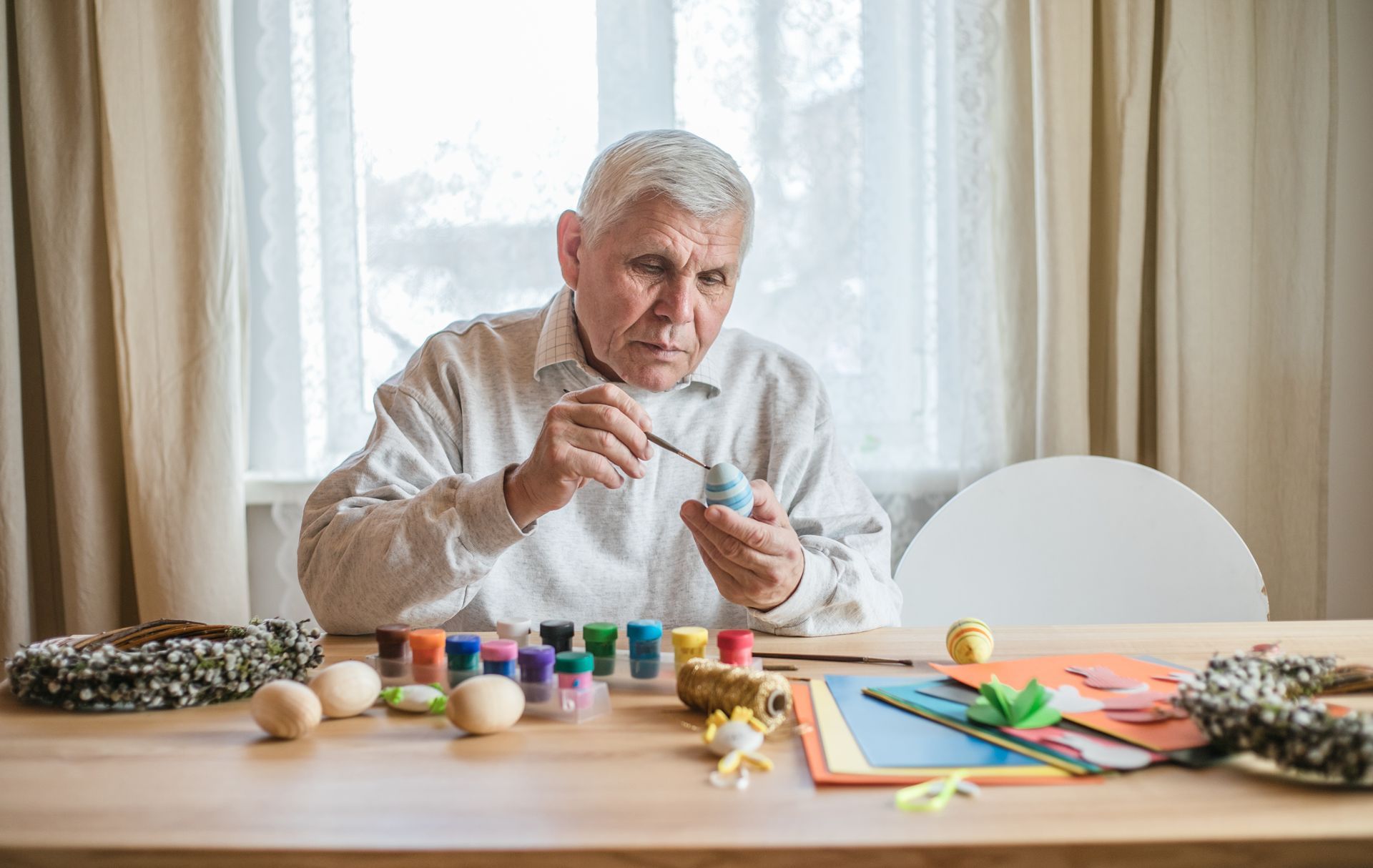 An elderly man is sitting at a table painting easter eggs.