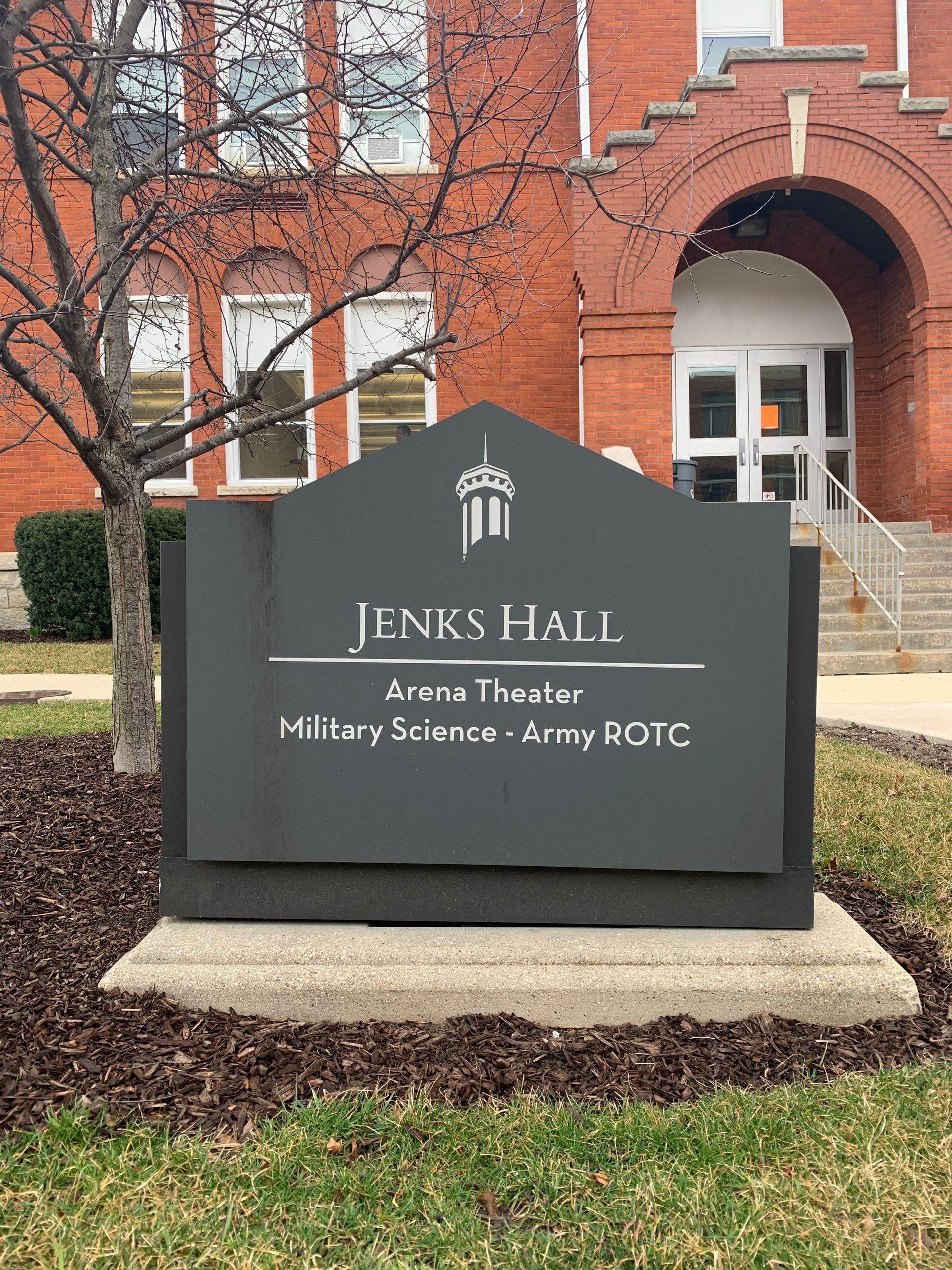 Sign for Jenks Hall, with details of arena theater and military science, in front of a brick building.