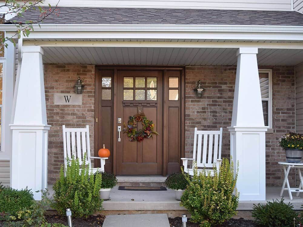The front porch of a brick house with two white rocking chairs and a wreath on the door