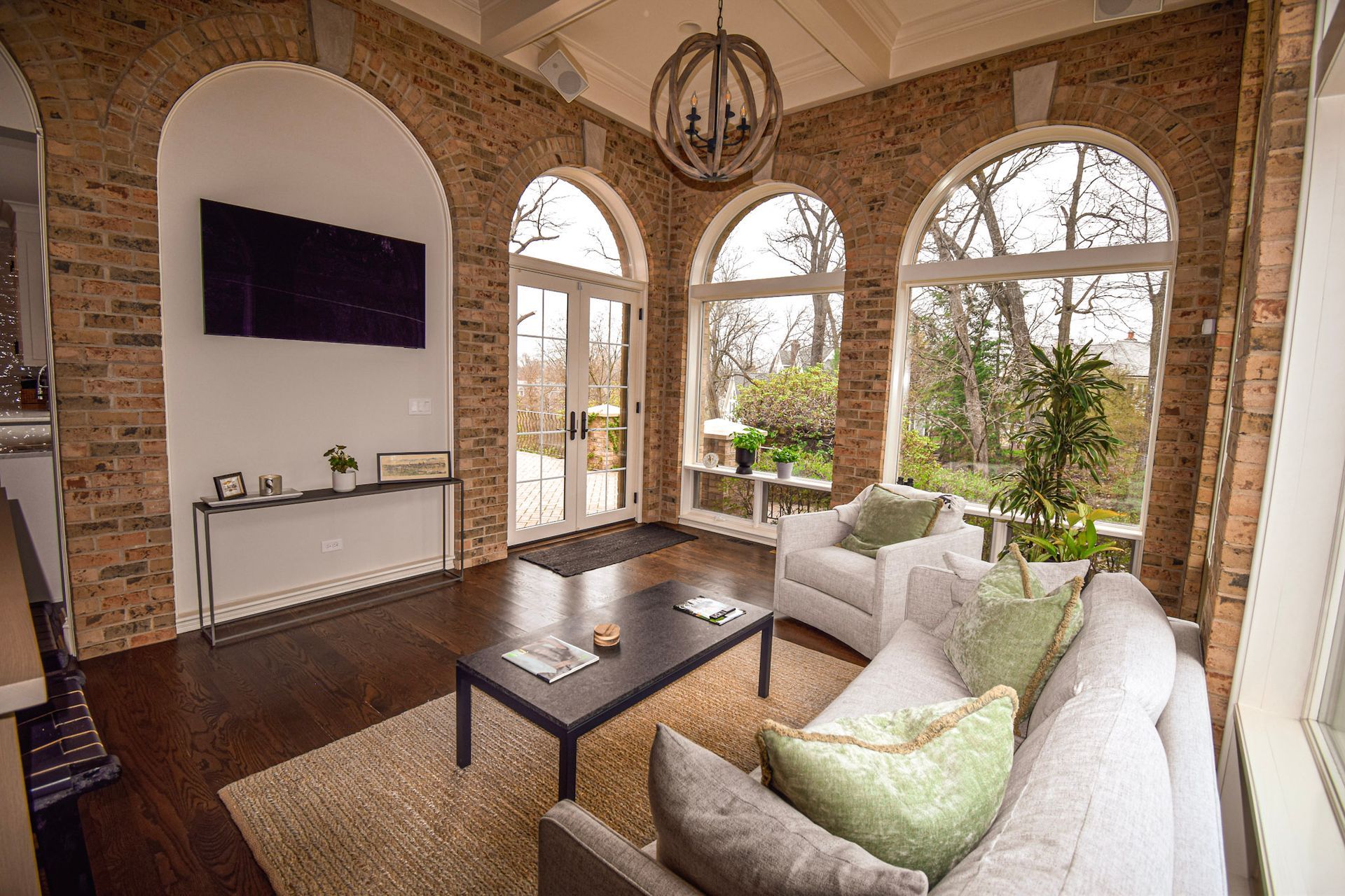Sunroom with brick arches, white furniture, large windows overlooking trees, and a mounted TV.