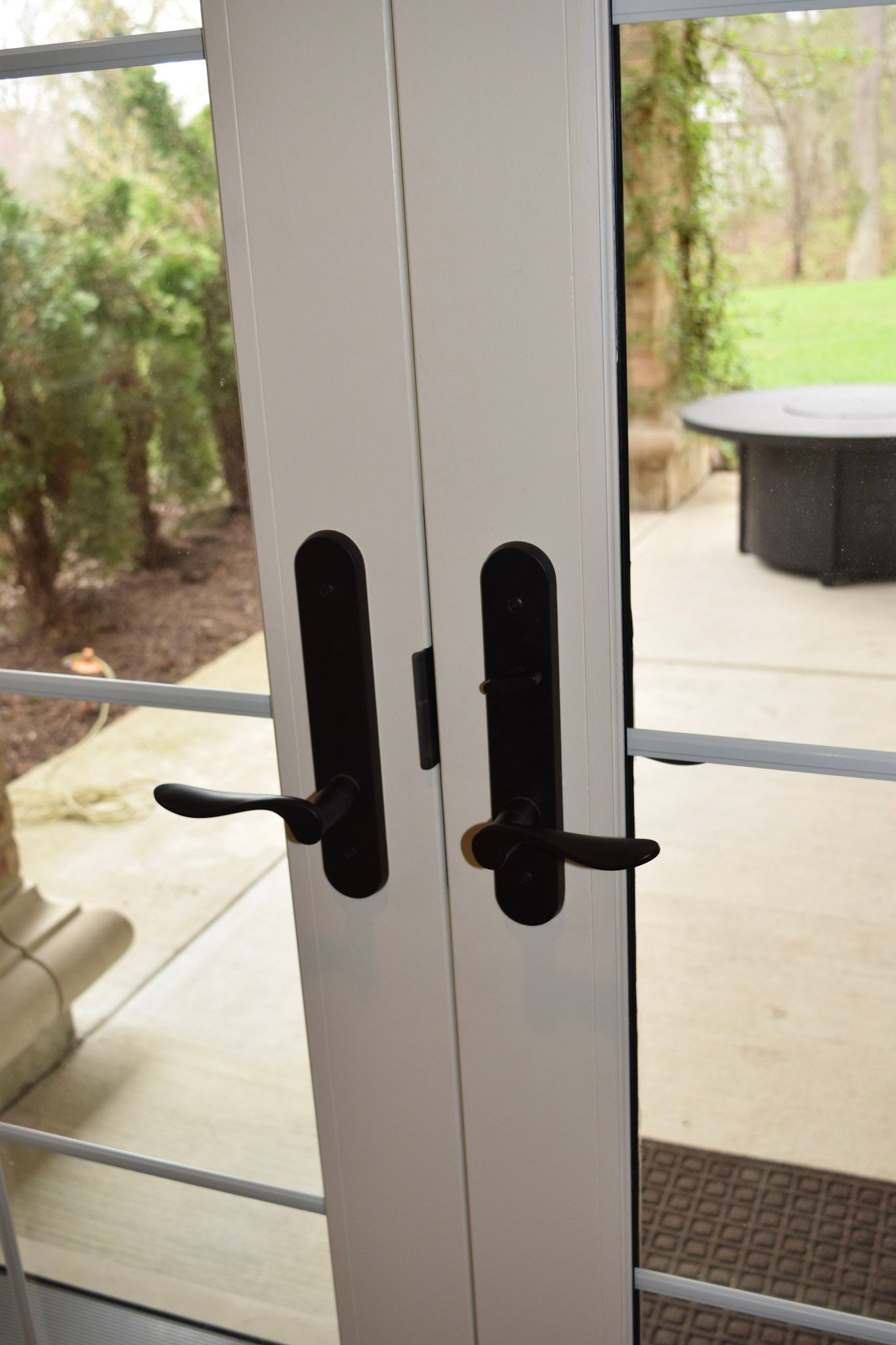 White double doors with black handles, open onto a patio with a table and foliage.