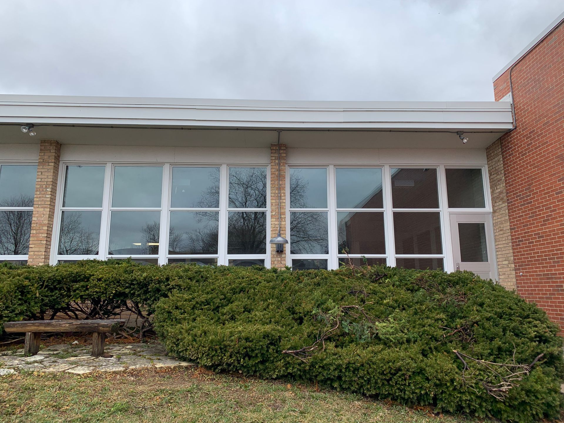 Building exterior with large windows, brick walls, and a hedge in front, under a cloudy sky.