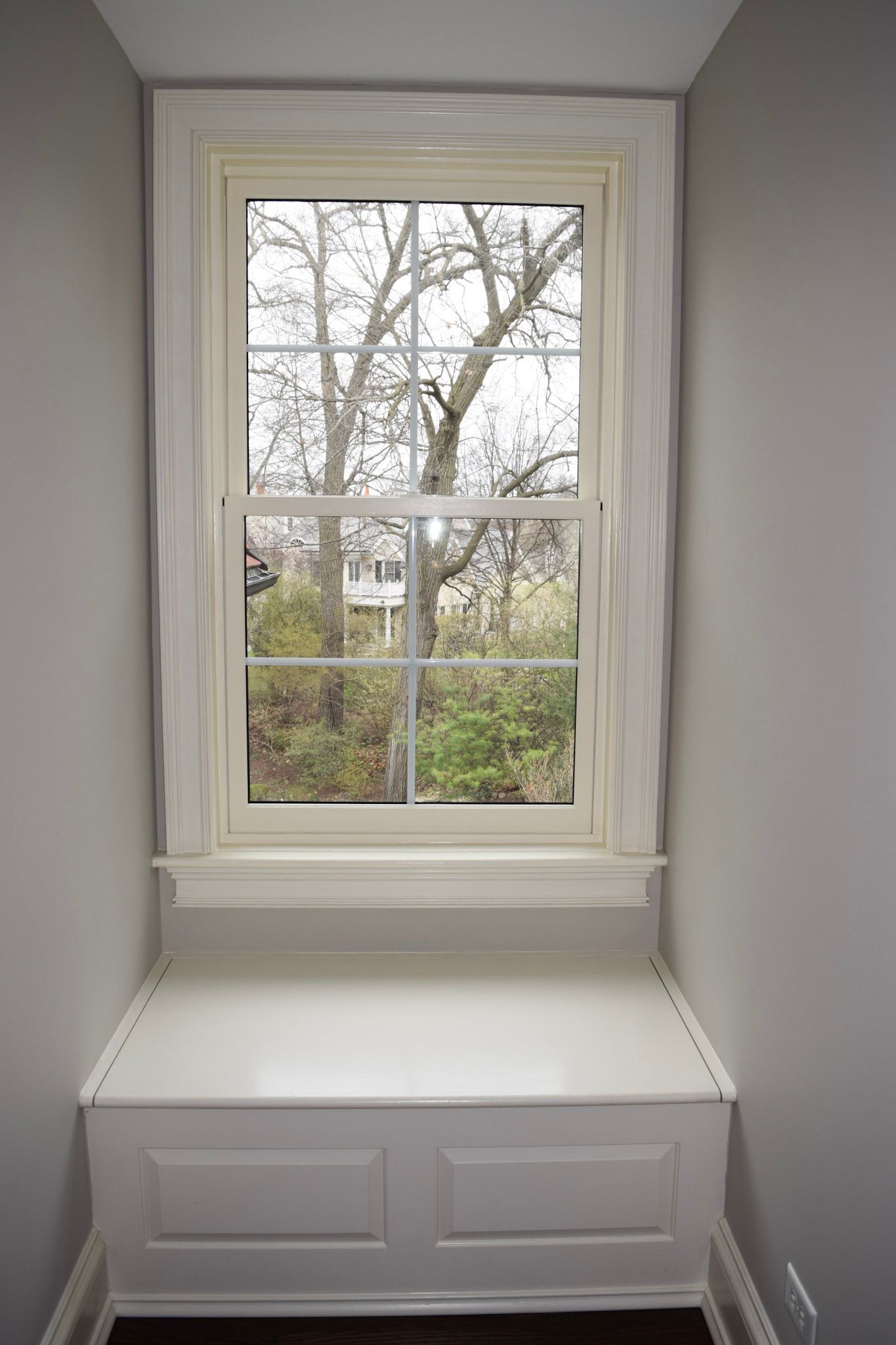 White window with built-in bench in a gray alcove, overlooking a tree-filled yard.