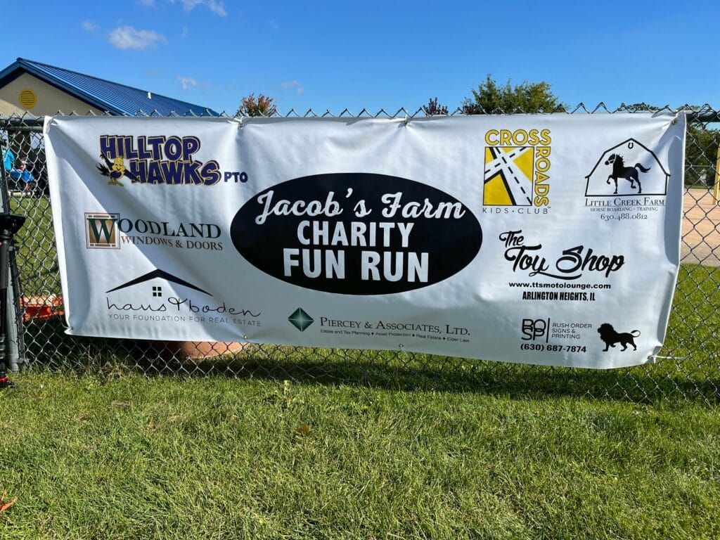 A banner for jacob 's farm charity fun run is hanging on a chain link fence.