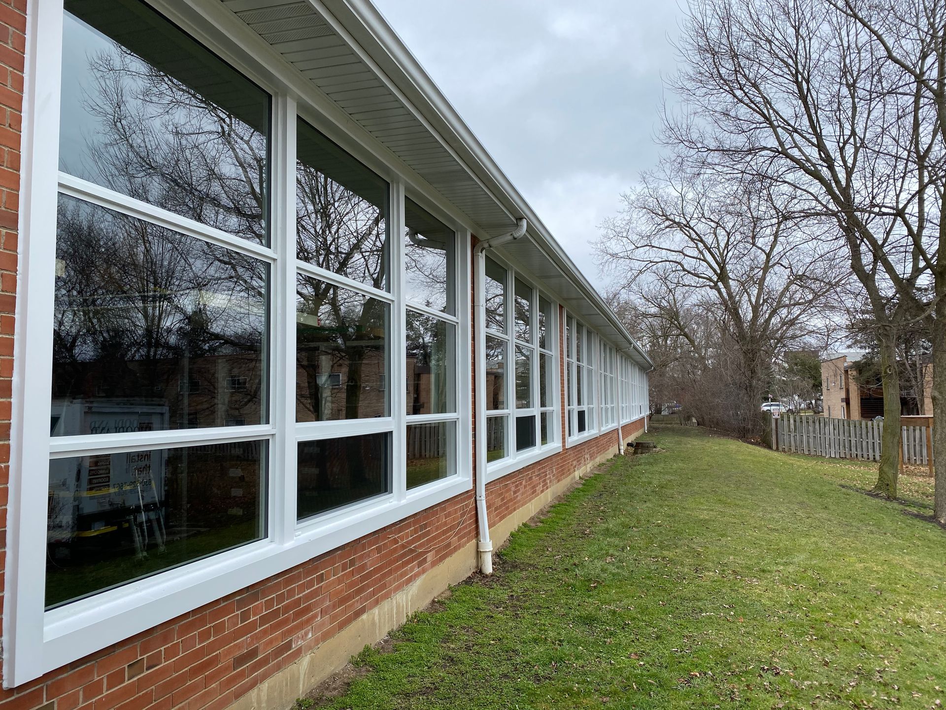 Long row of windows on a brick building with white frames, overlooking a grassy area and trees on a cloudy day.