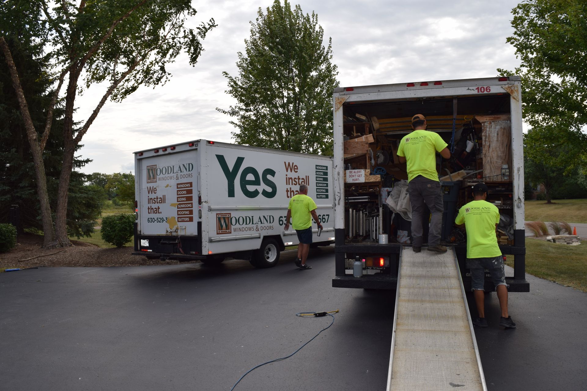 Movers in green shirts loading furniture into a truck with a ramp on a driveway next to another truck.