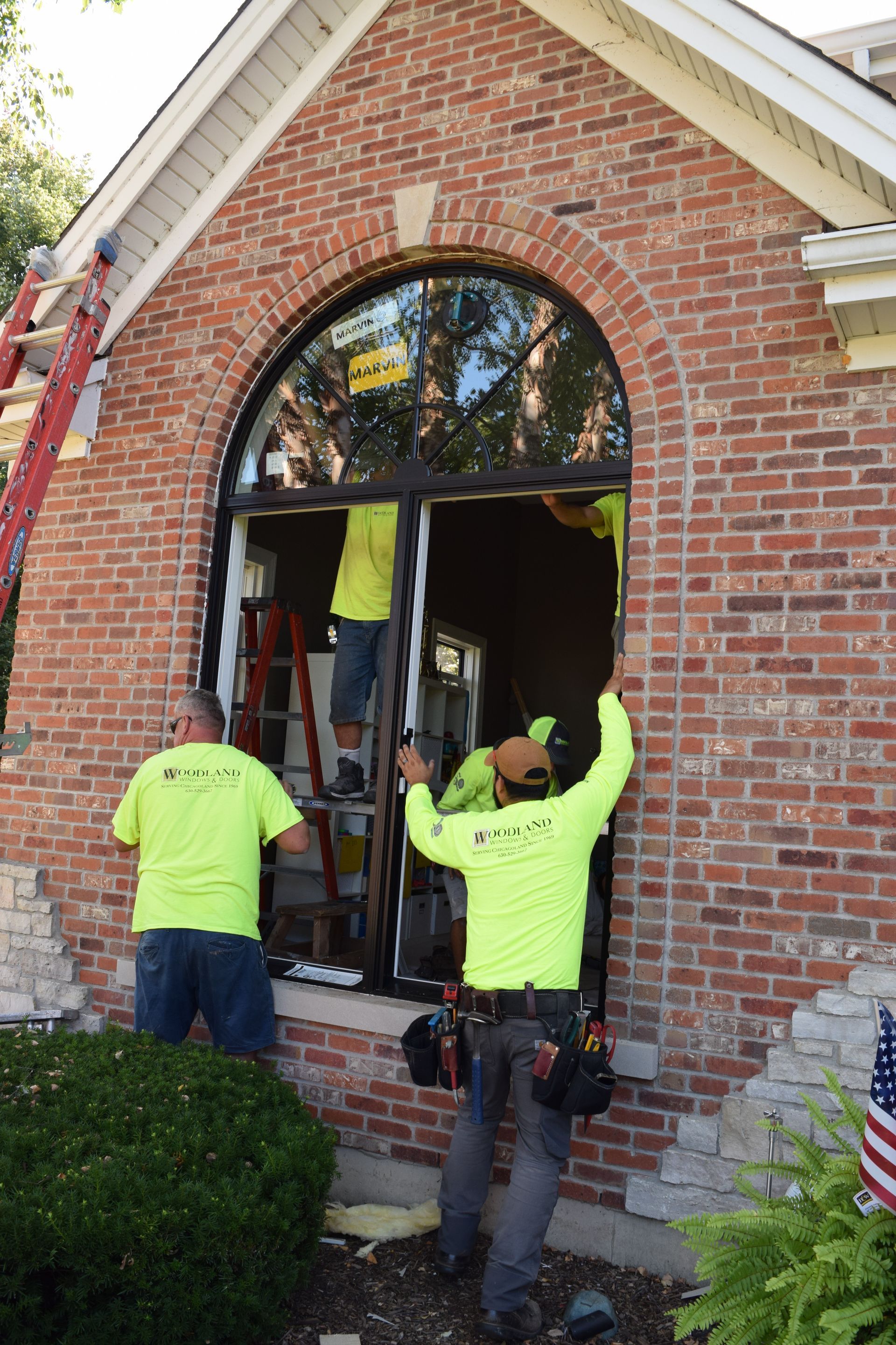 Construction workers installing an arched window in a brick building. They wear yellow shirts and work outside.
