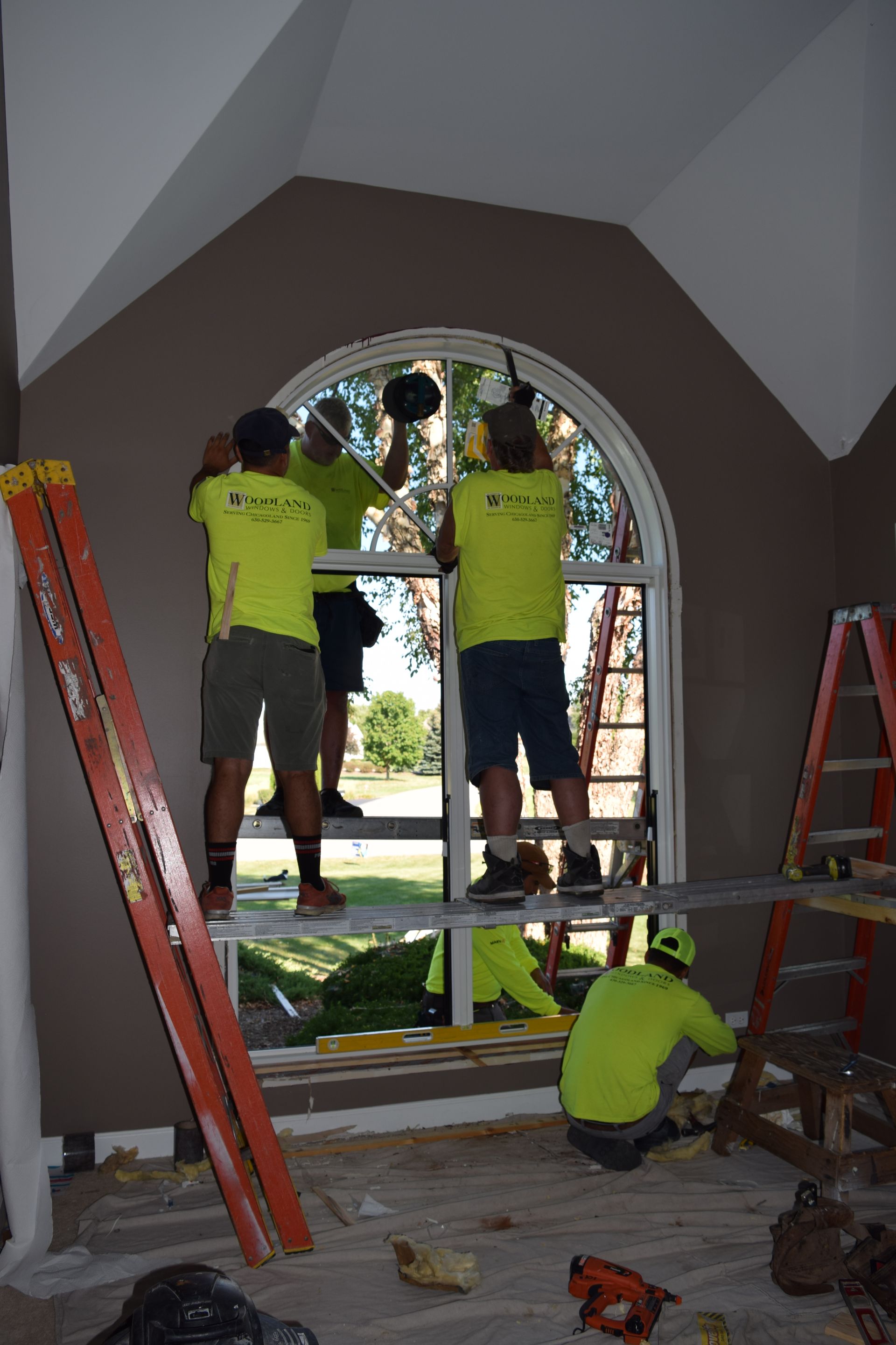Four workers installing a large arched window, two on a scaffold, one on ground, inside a room.
