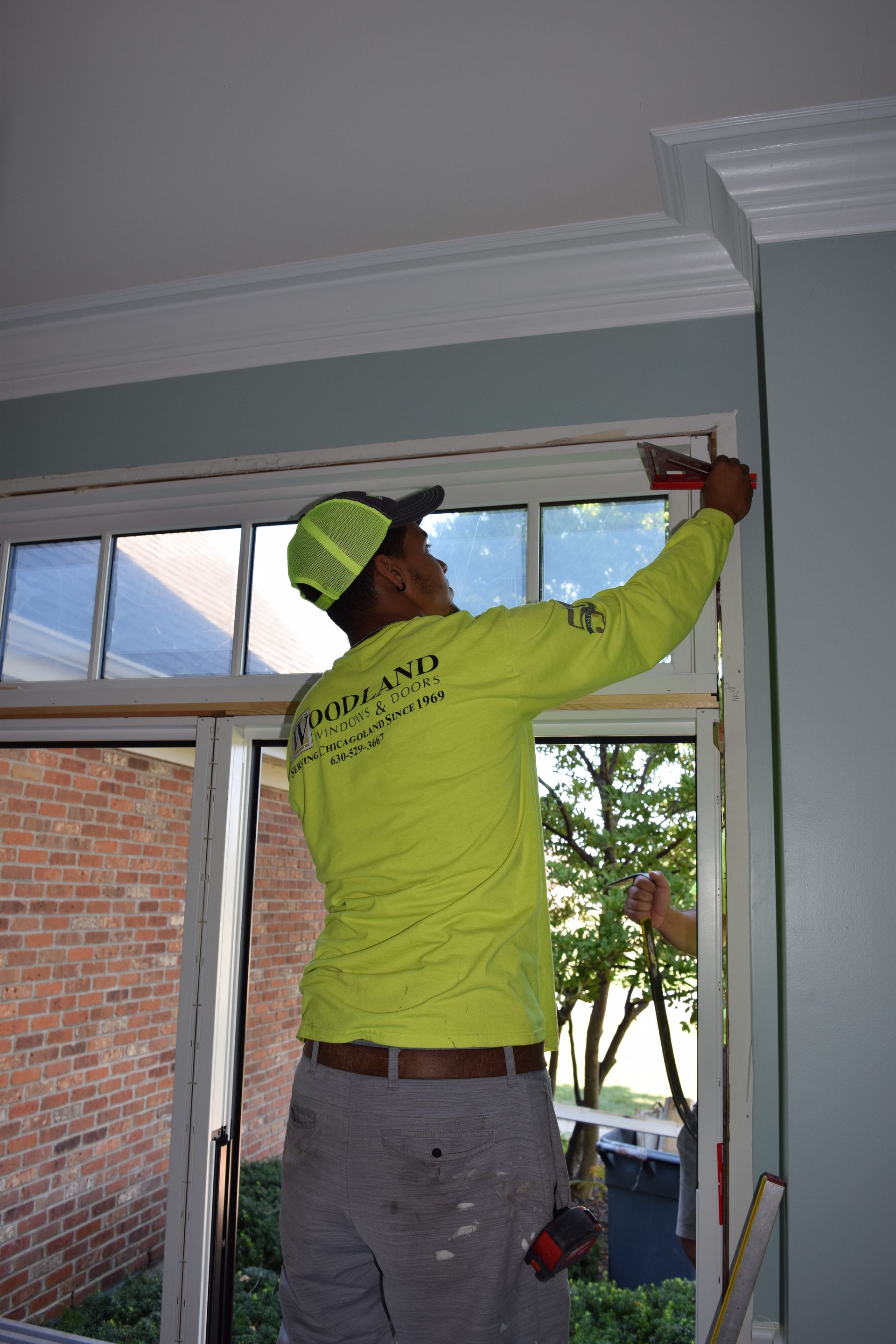 Man in yellow shirt installing trim around a window. Interior shot with brick building visible outside.