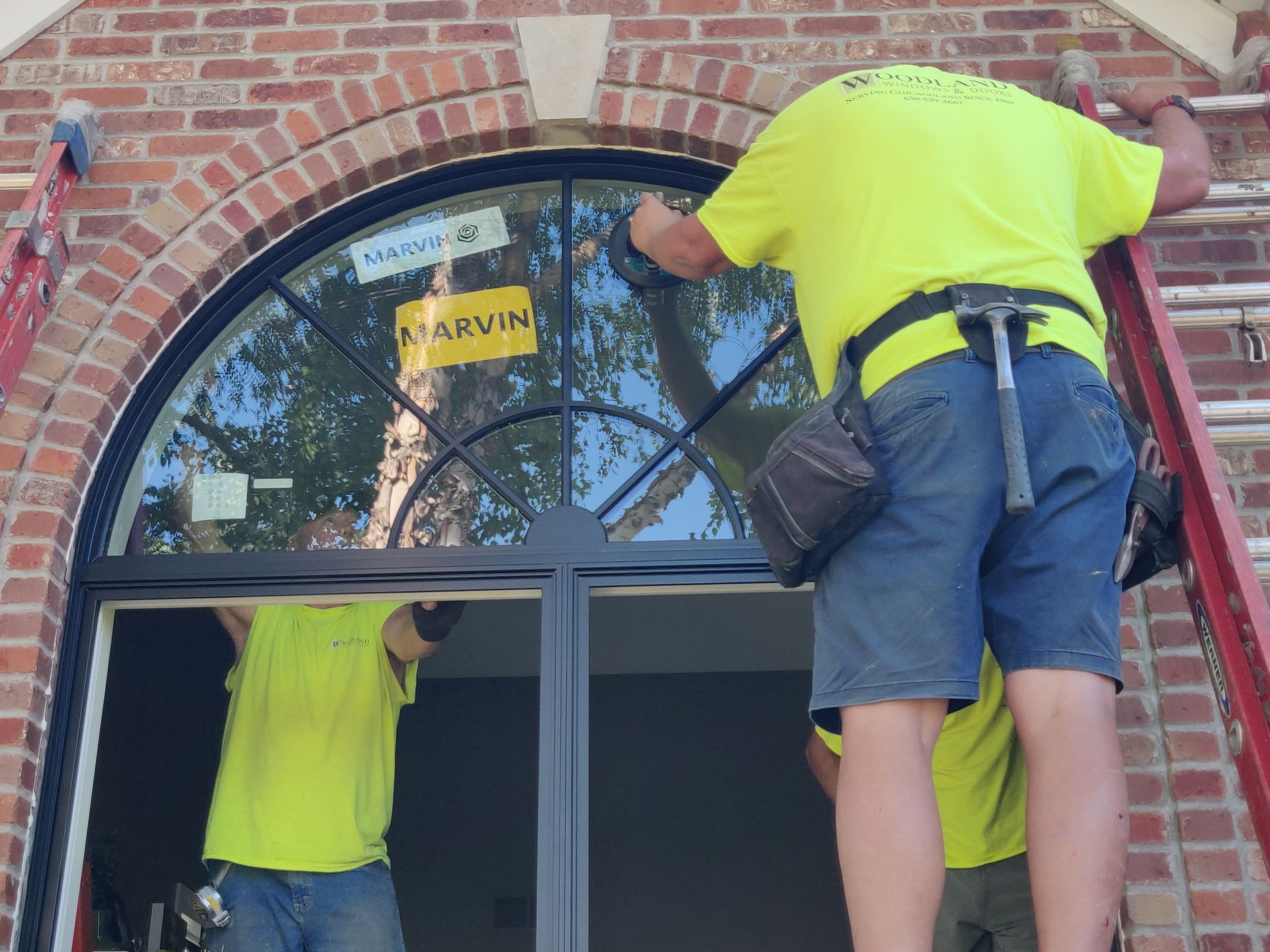 Workers installing a black arched window in a brick building. One person is on a ladder.