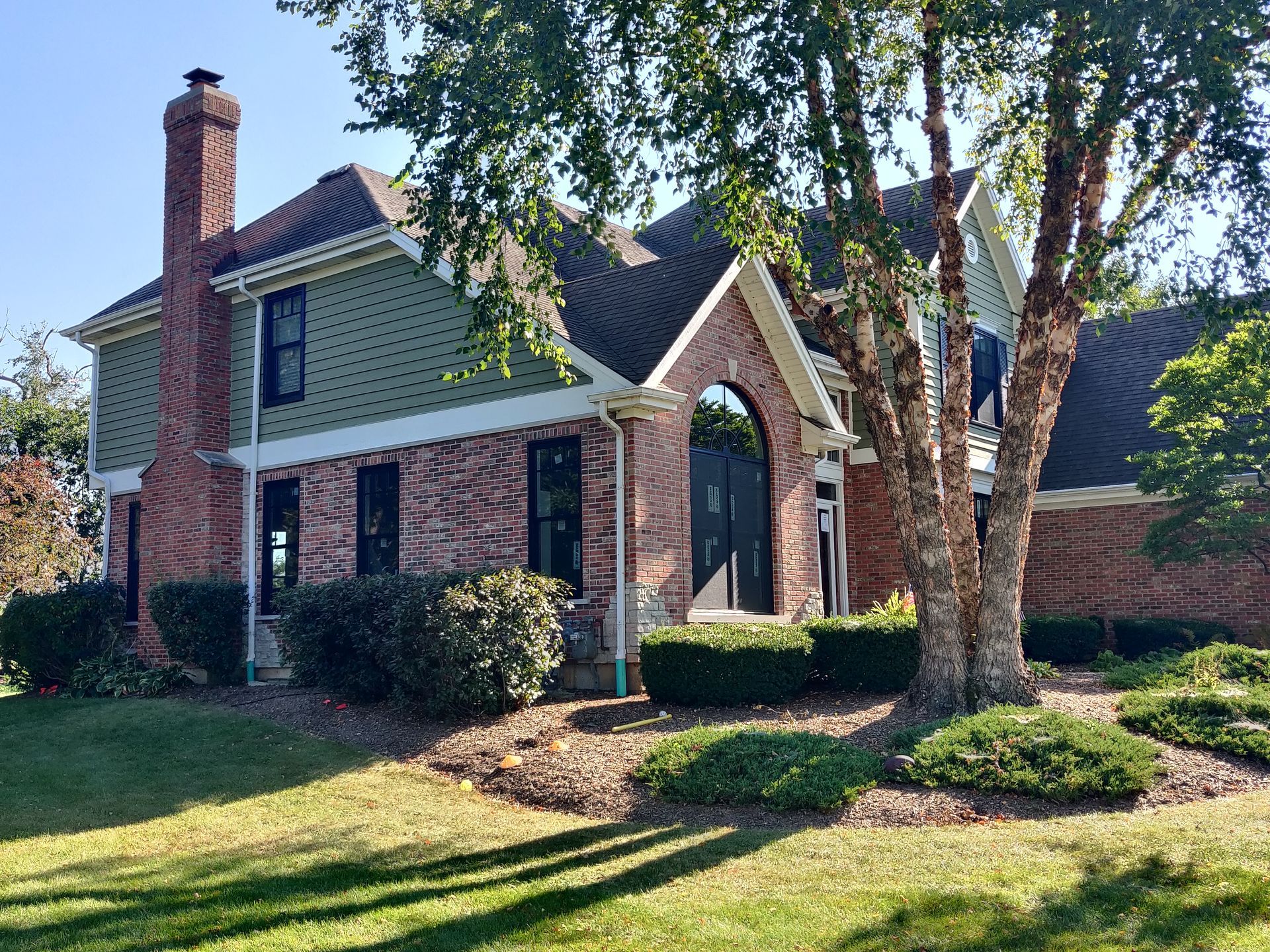 Two-story brick house with a green upper level and large windows, tree in front, sunny day.