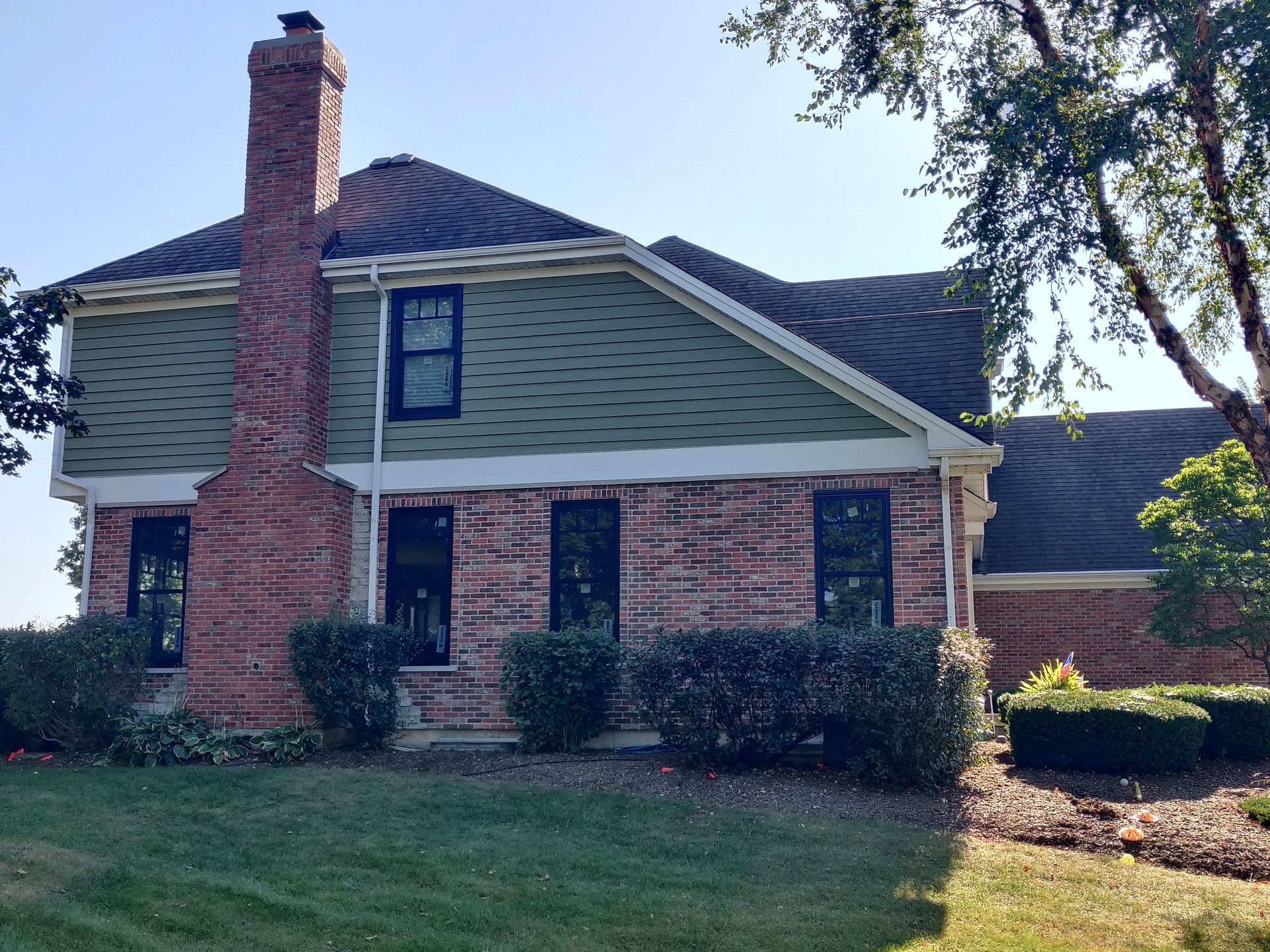 Brick house with green siding, black-framed windows, and a tall brick chimney, on a sunny day.