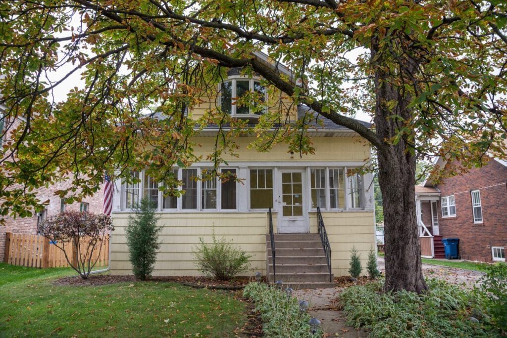 A yellow house with a porch and stairs is surrounded by trees.