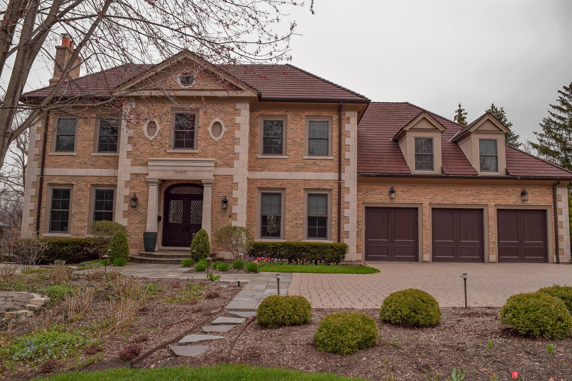 Large brick house with brown roof and garage; stone walkway and bushes in front.