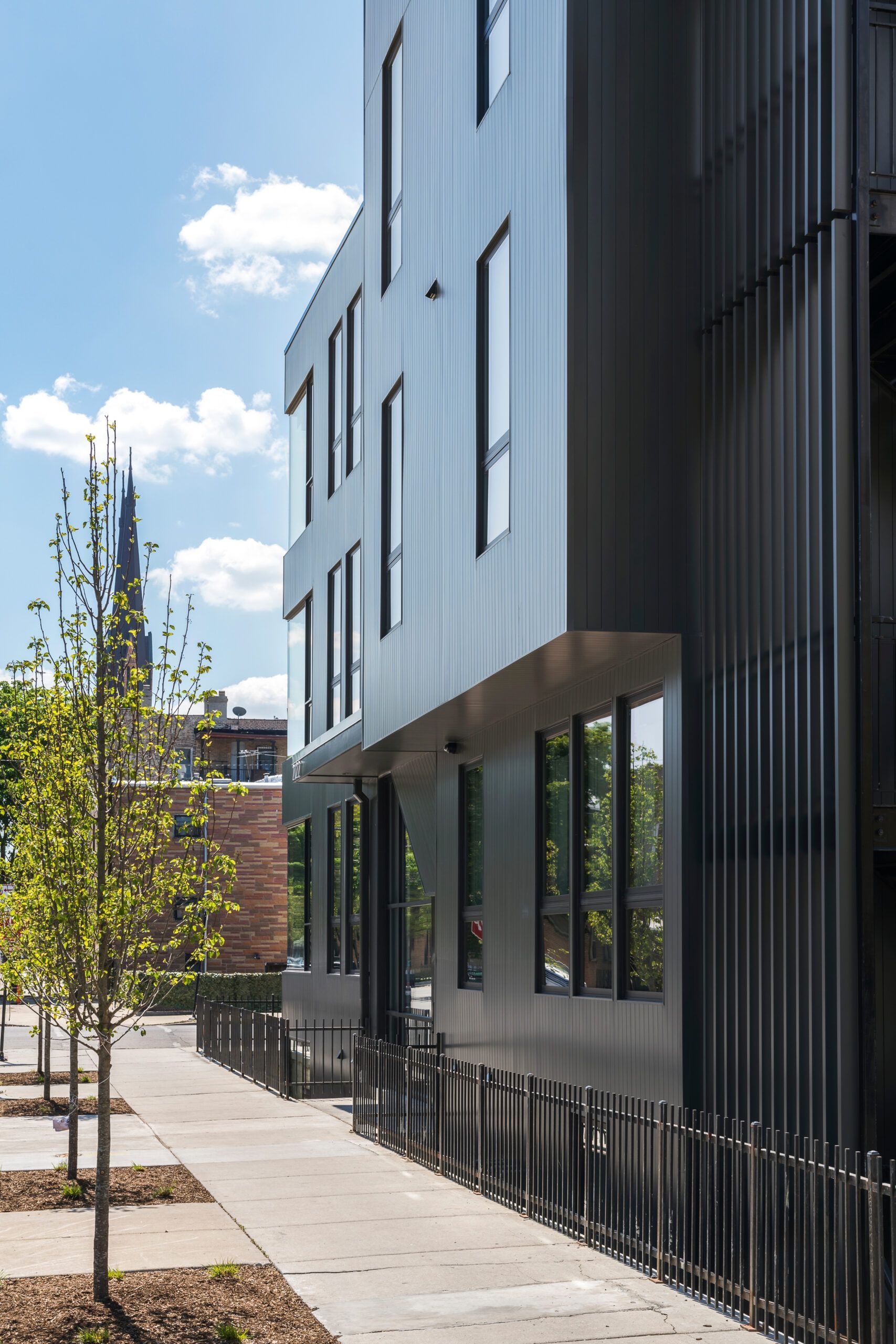 Modern black building with vertical windows, a sidewalk, and trees. Blue sky with clouds.