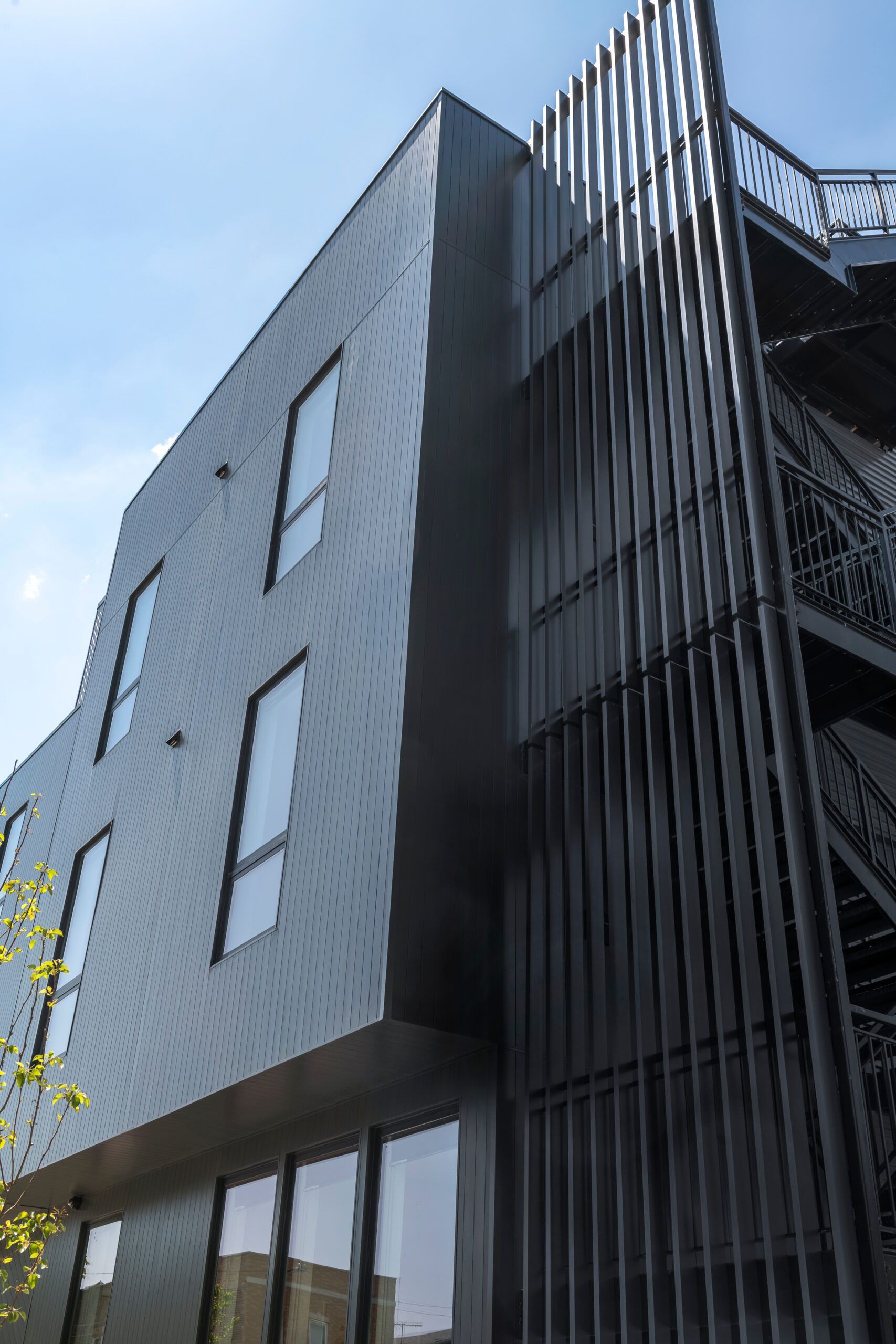 Dark gray modern building with vertical black accents, outdoor staircase, and a bright blue sky.
