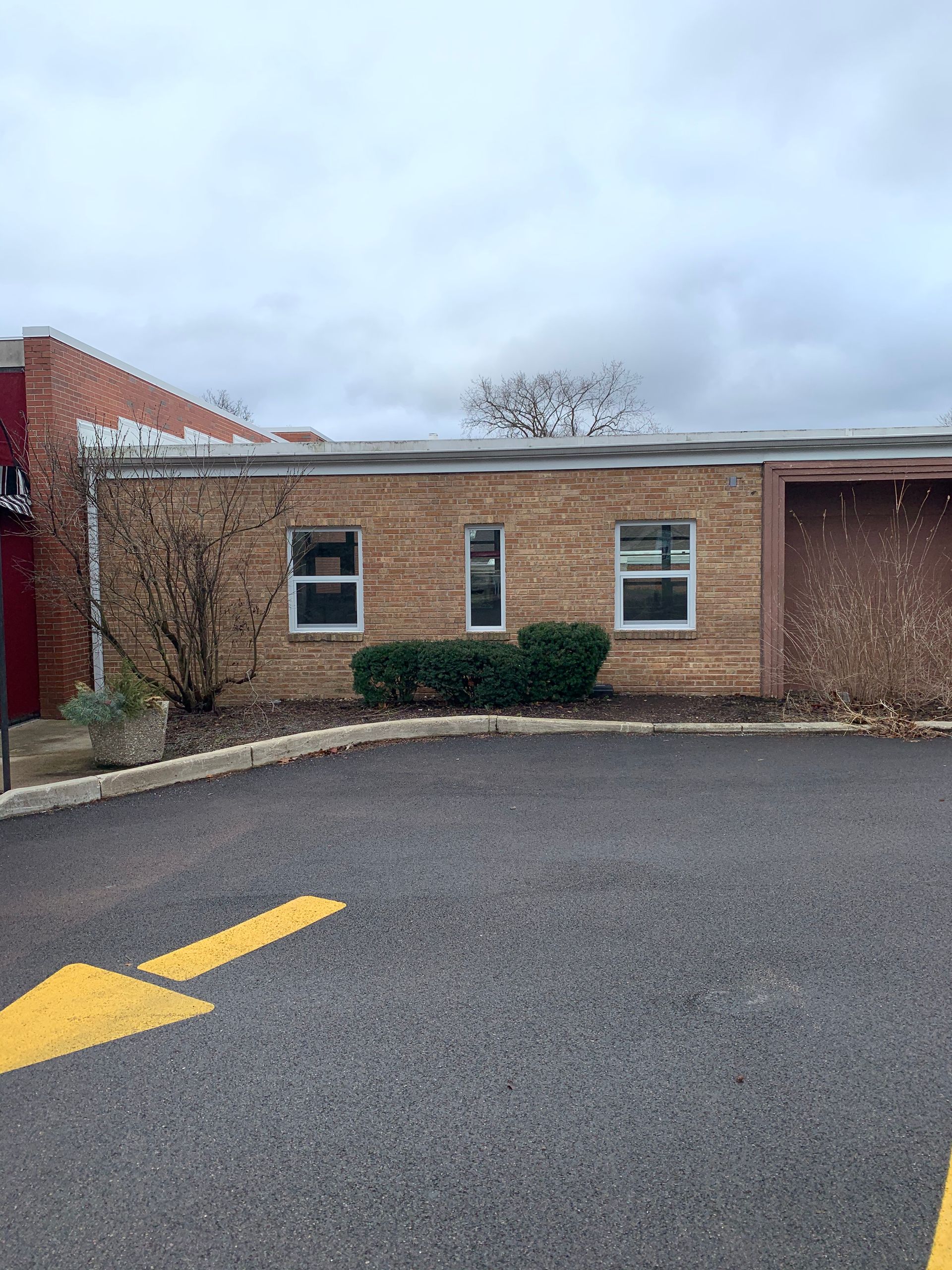 Brick building with three windows and a yellow arrow on the asphalt in front. Overcast day.