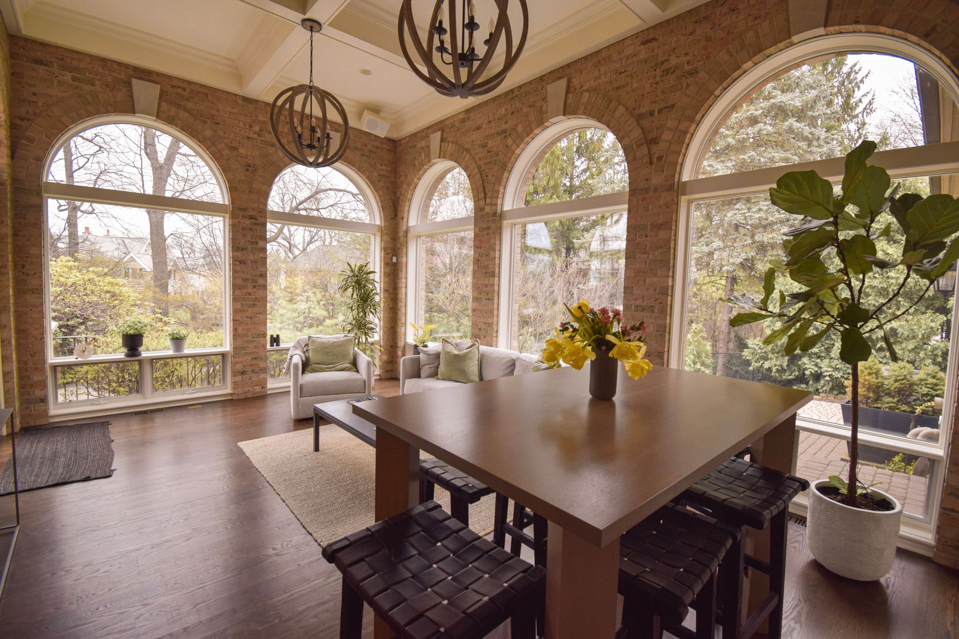 Sunroom with arched windows, brick walls, dining table, and lush greenery outside.
