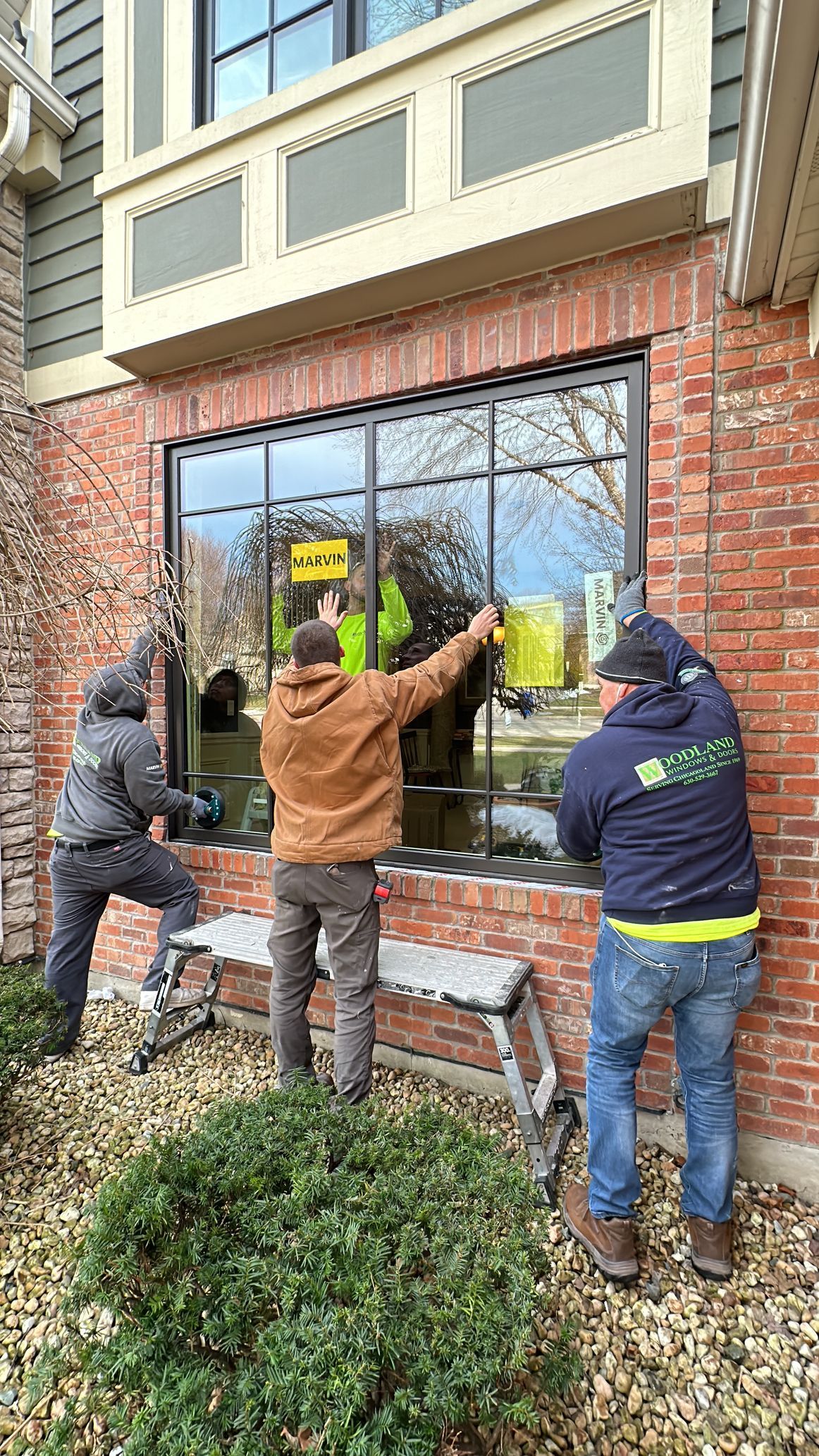 Three workers installing a large black-framed window in a brick building. One is on a bench.