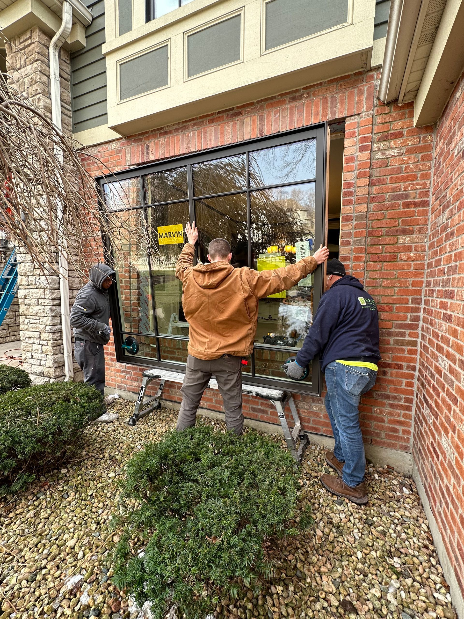 Three workers installing a large black framed window in a brick building.