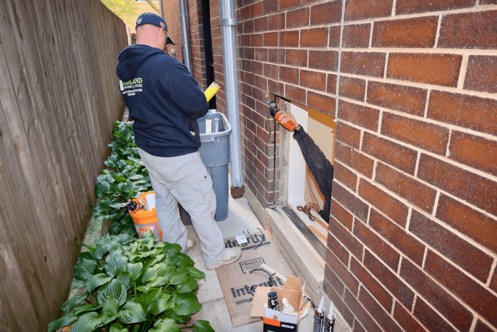 A man is working on installing a window on the side of a brick building.