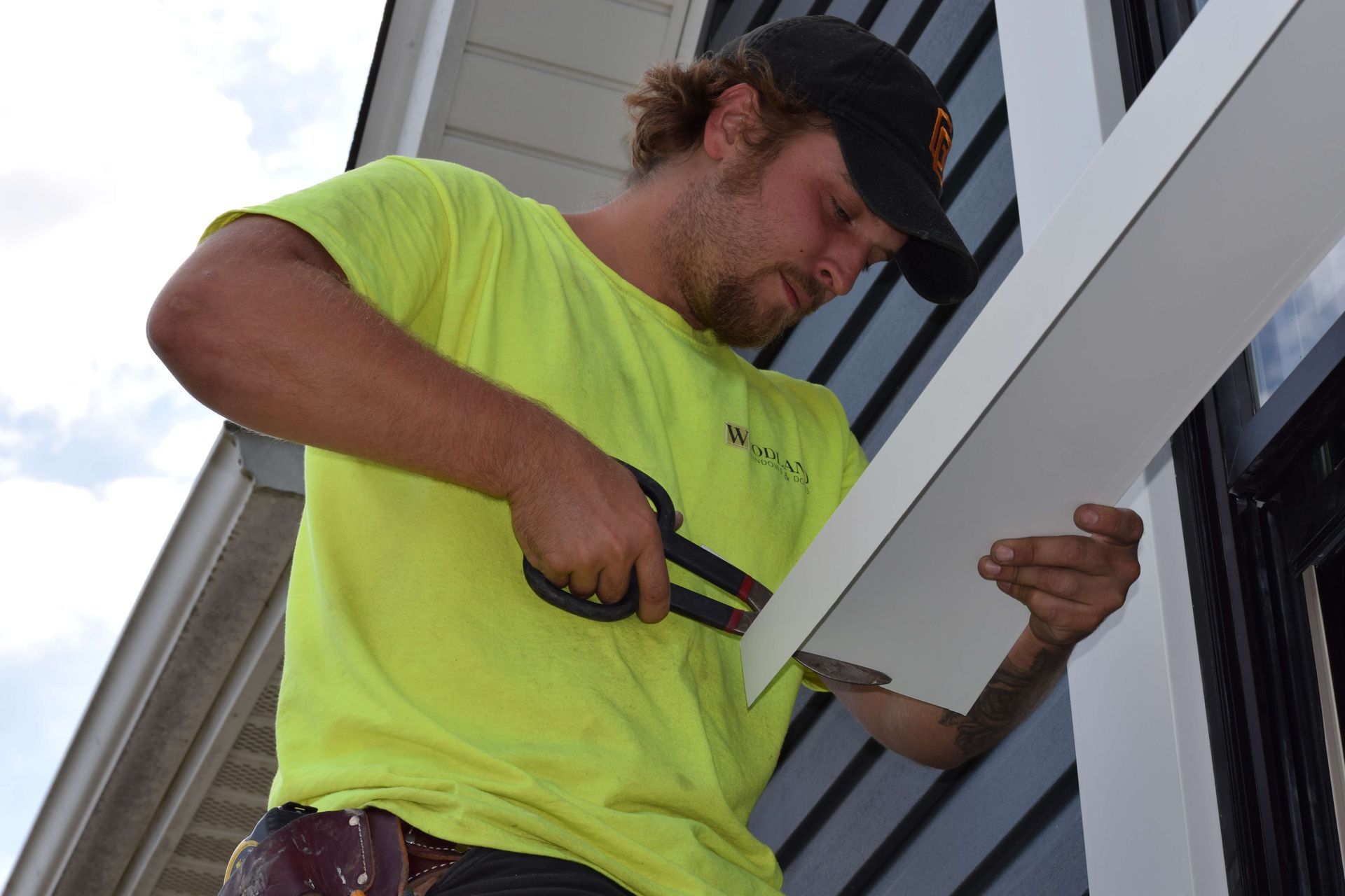 A man in a yellow shirt is cutting a piece of siding with a pair of scissors.