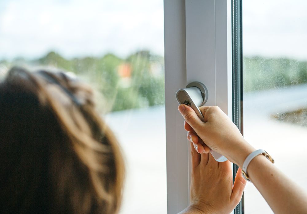 Close-up of a person's hand turning a white window handle to open a window, with a blurred outdoor background.