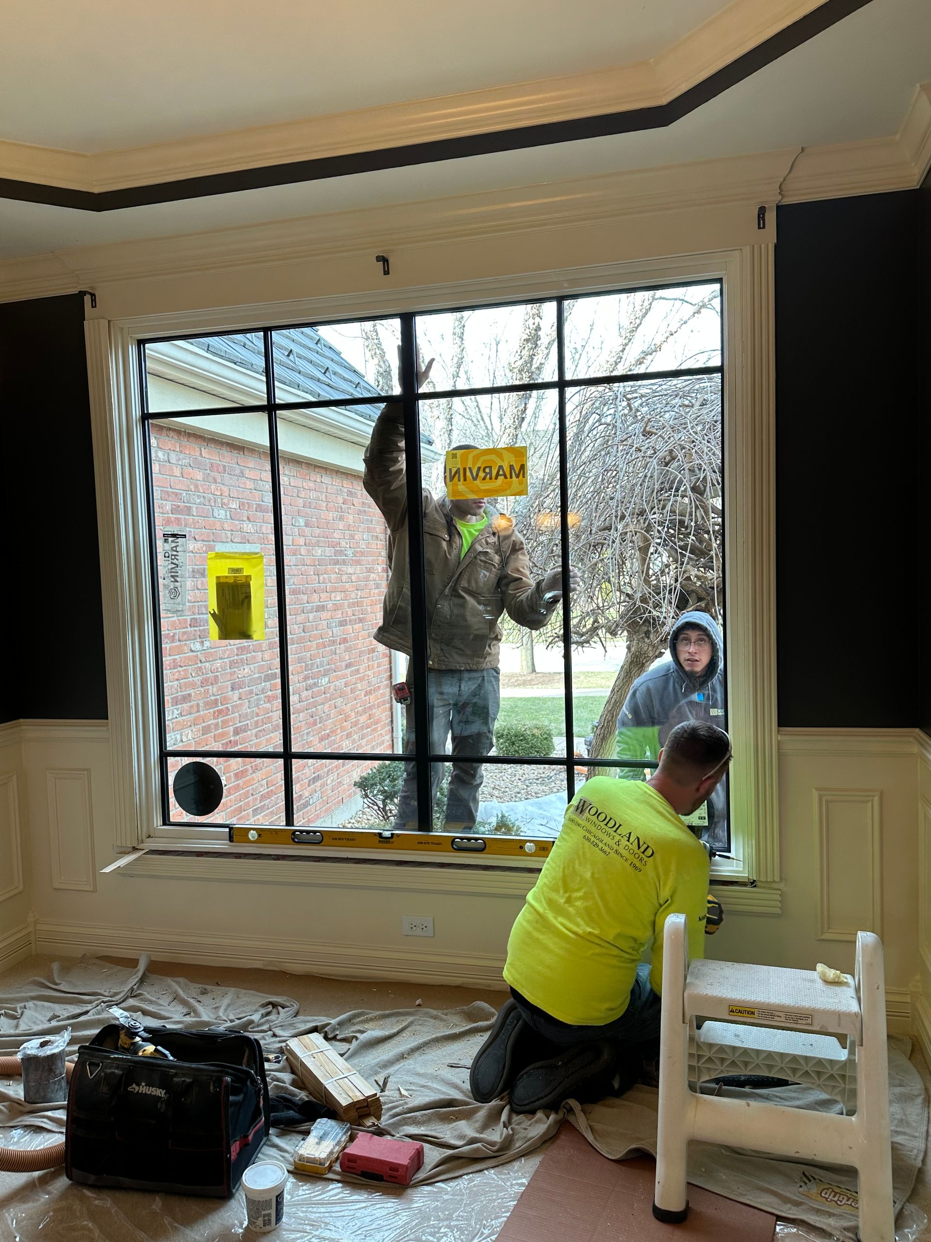 Workers installing a large window with grid. One kneels, two inside. Exterior view, tools and materials on floor.