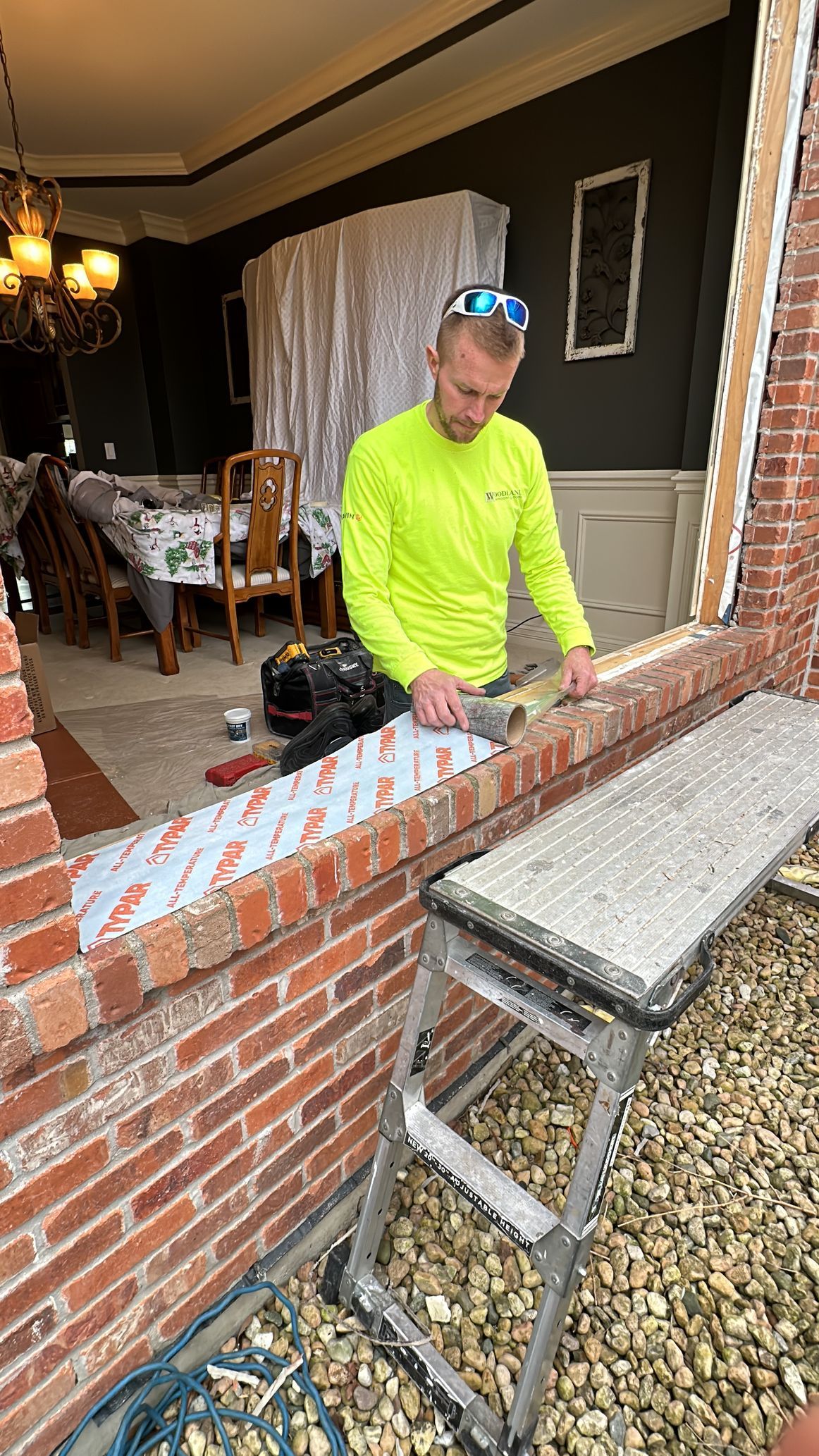 Man in neon shirt replacing a window frame, working on a brick exterior with a step stool.