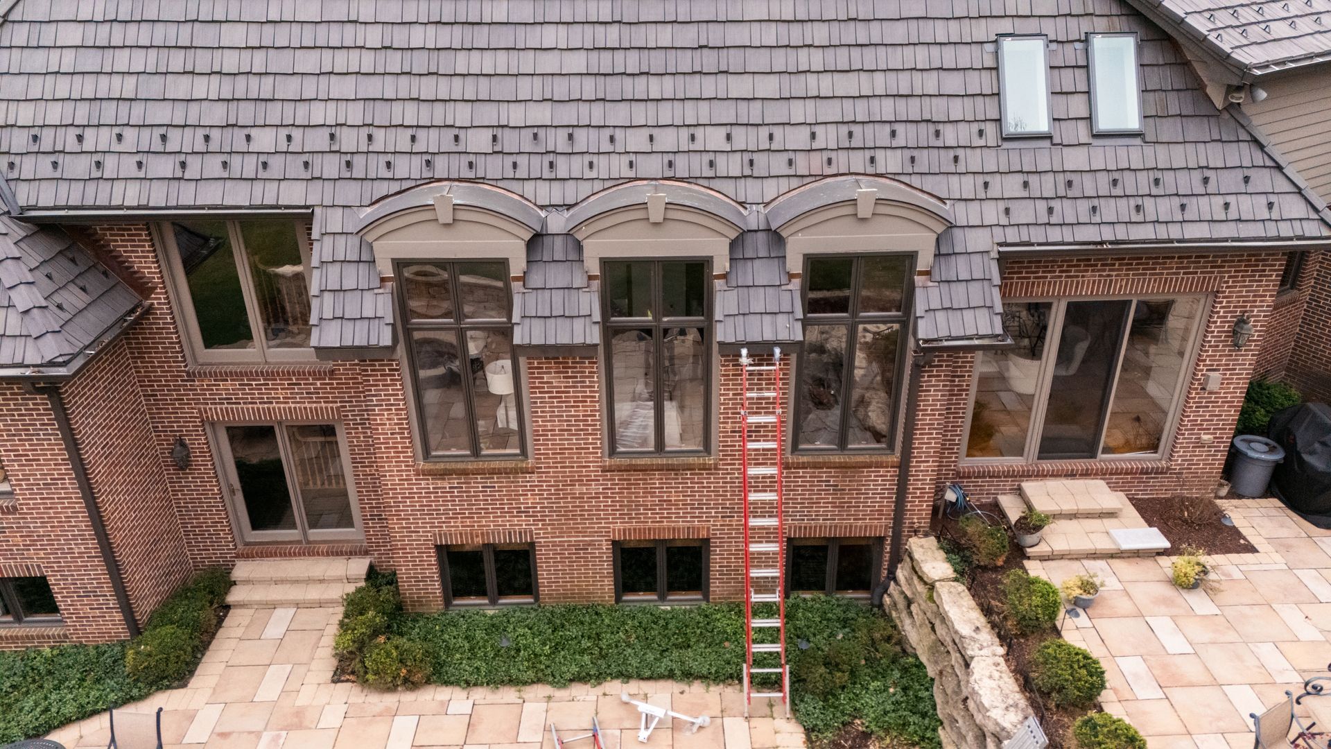 Red brick house exterior with windows, a ladder, and a stone patio.
