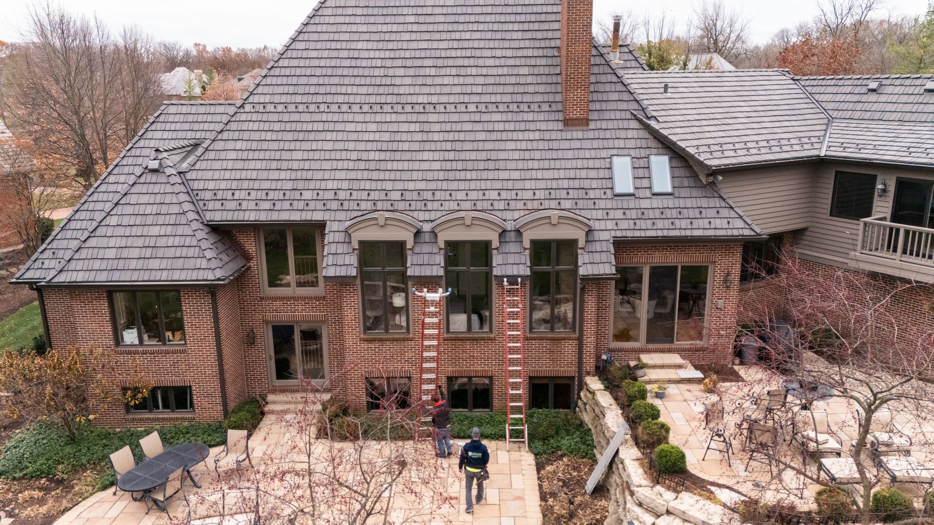 Large brick house with gray shingle roof, patio, and trees in the background. A person stands in front of the house.