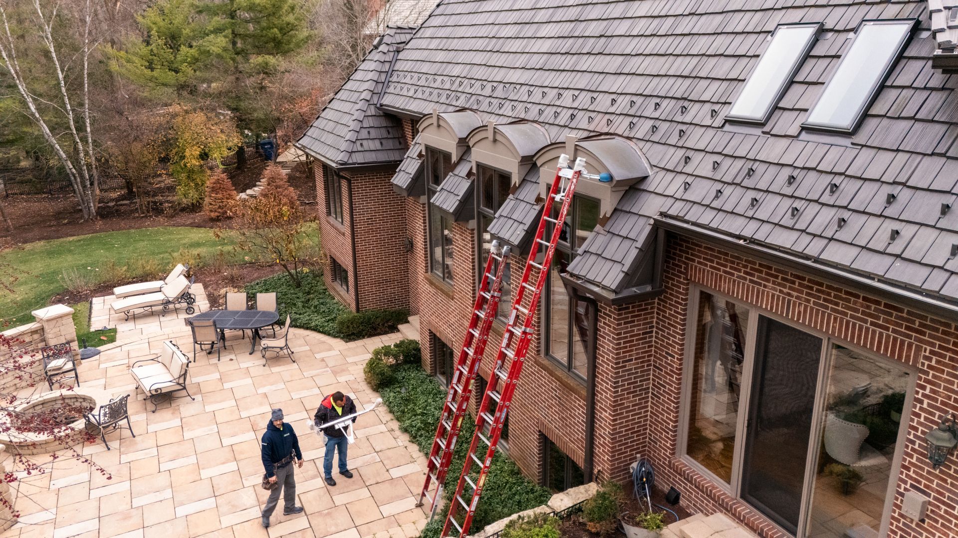 Workers on a roof repair, brick house with patio, fall landscape.