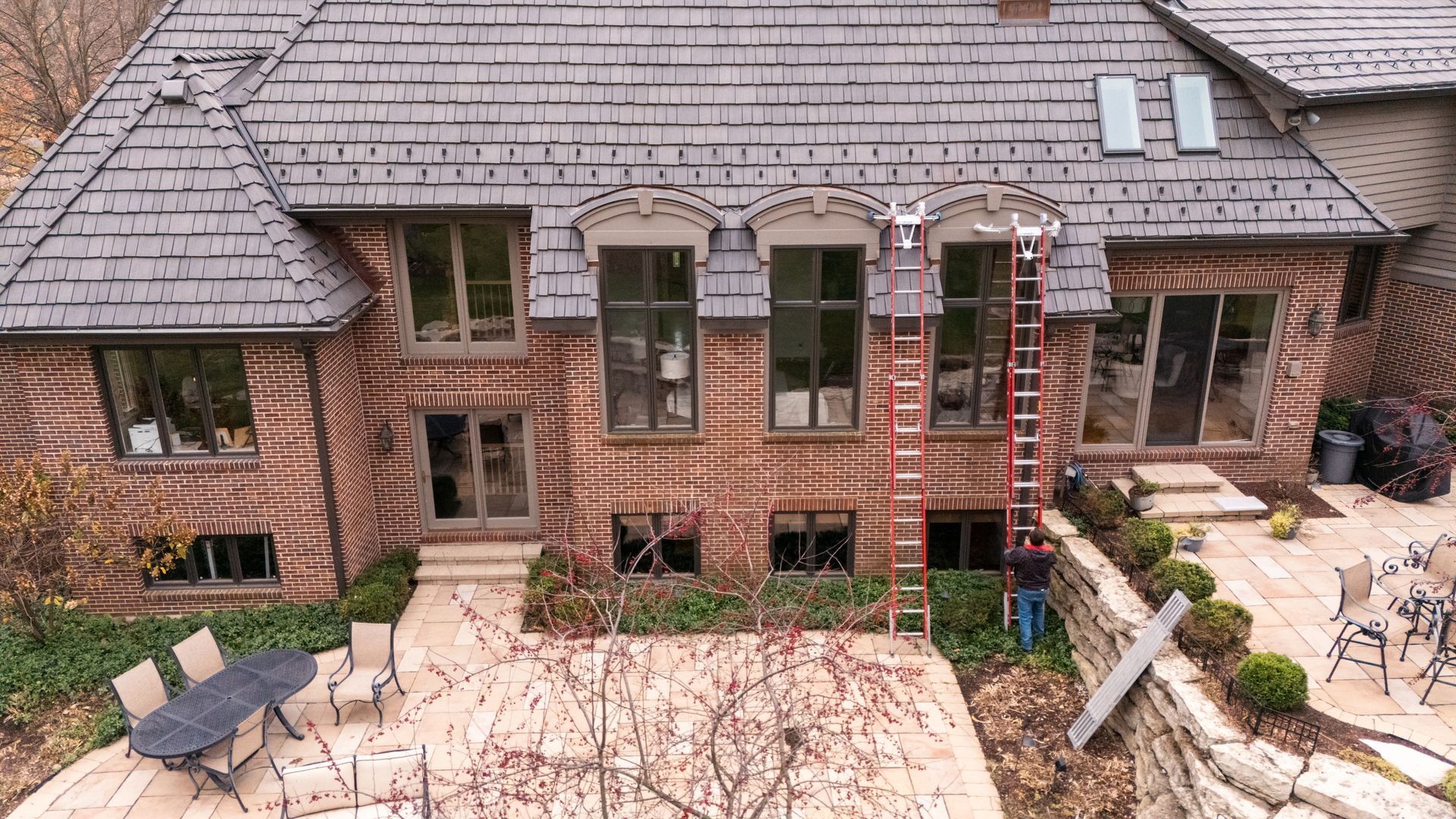 Brick house with dark roof, windows, and patio, ladder against the wall.