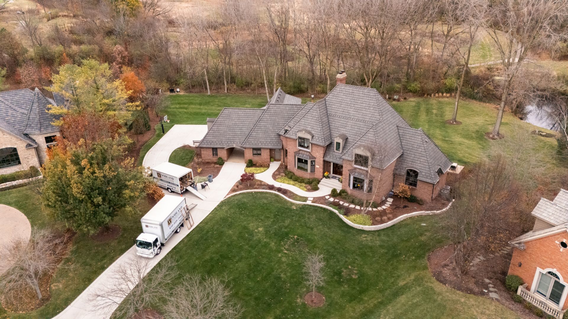 Two moving trucks parked at a large brick house with a curved driveway and green lawn.