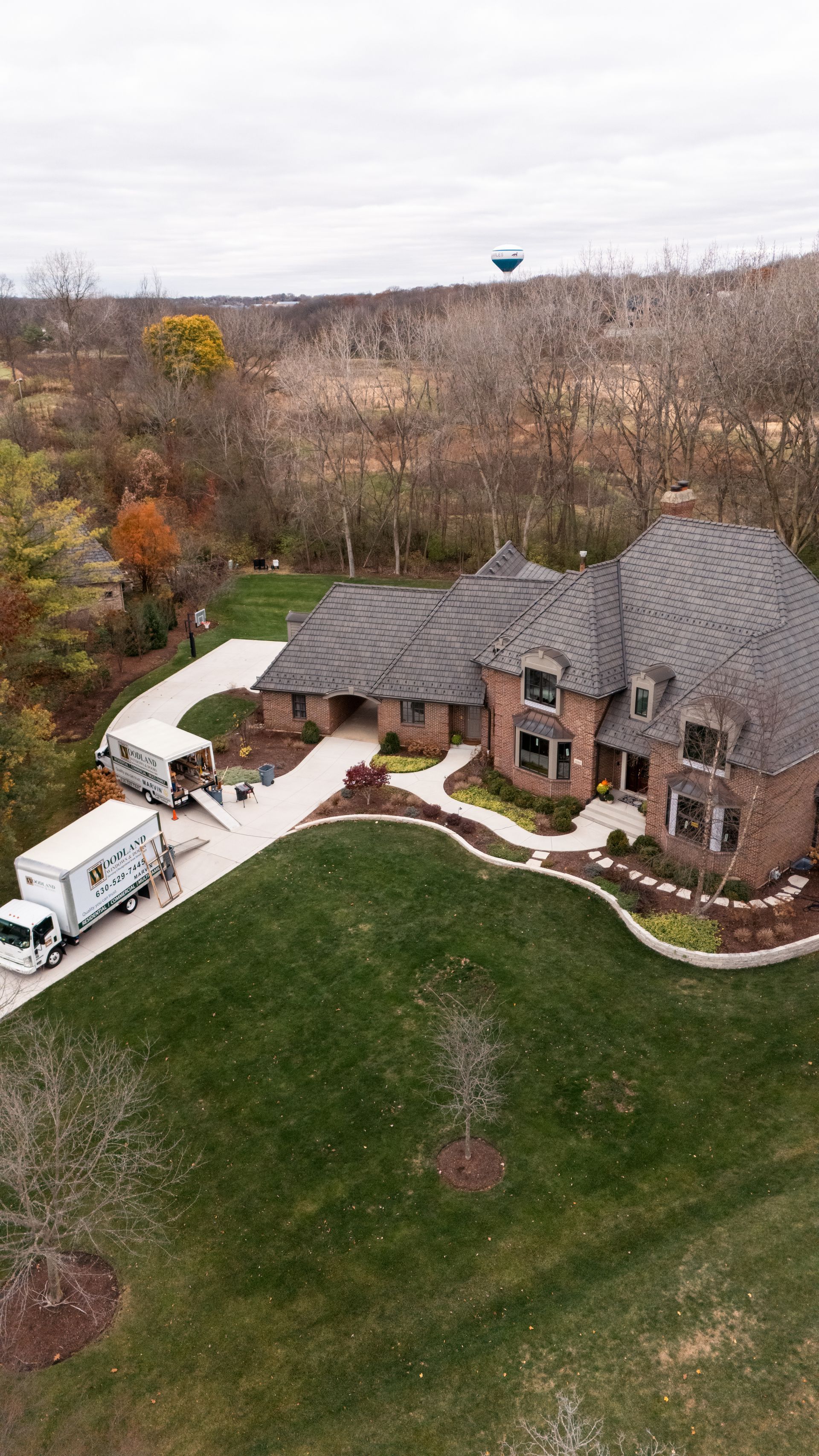Moving trucks parked in front of a large brick house on a green lawn, with trees in the background.