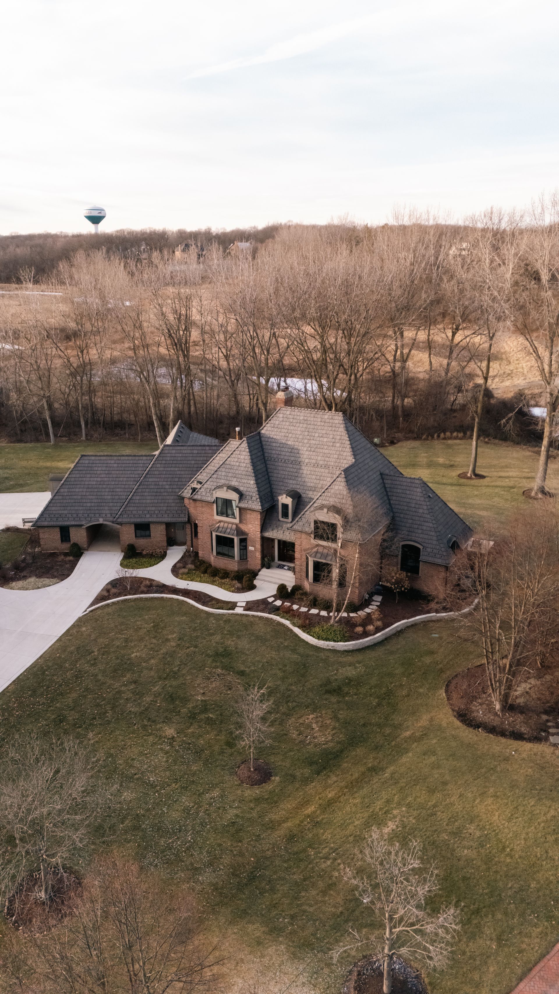 Brick house with dark roof on a grassy lawn, with a driveway leading to it. Trees and a water tower in the background.