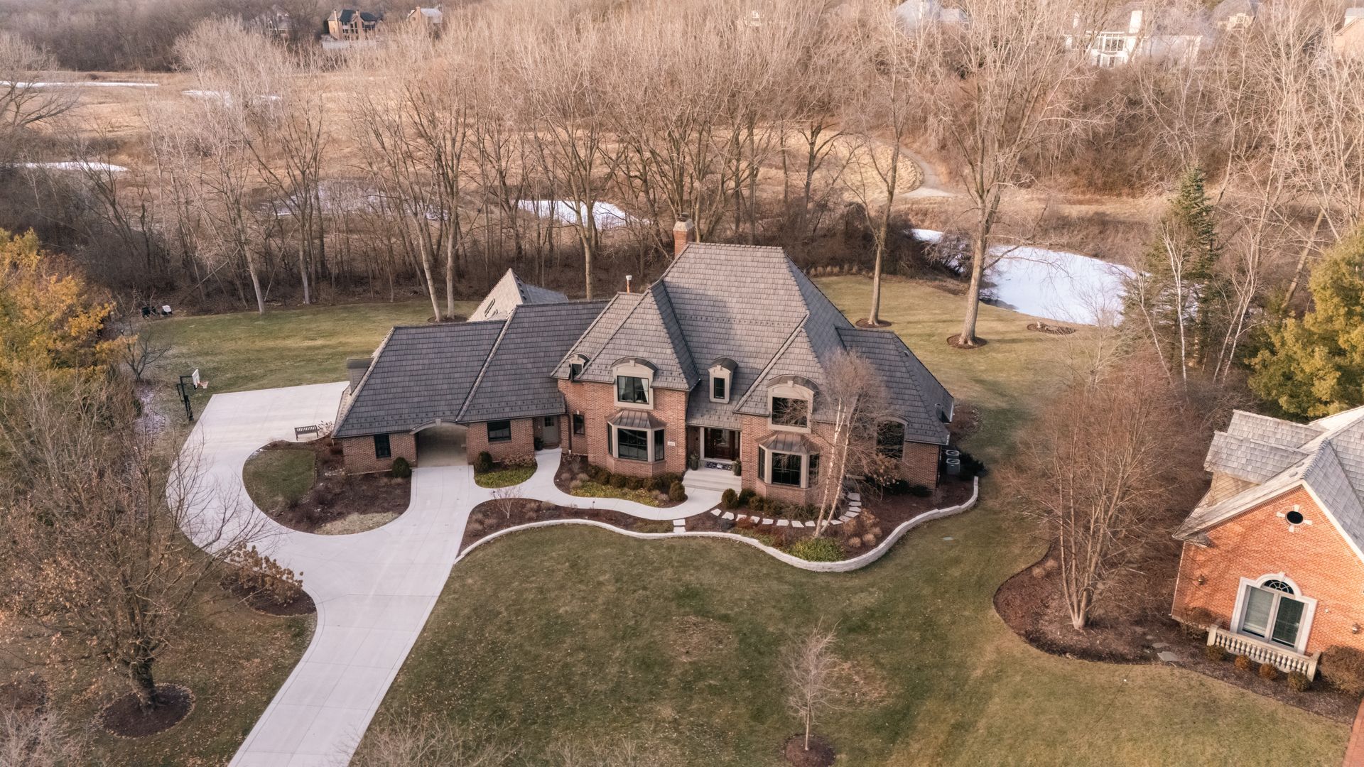 Large brick house with dark roof and long driveway, set in a grassy yard with trees.