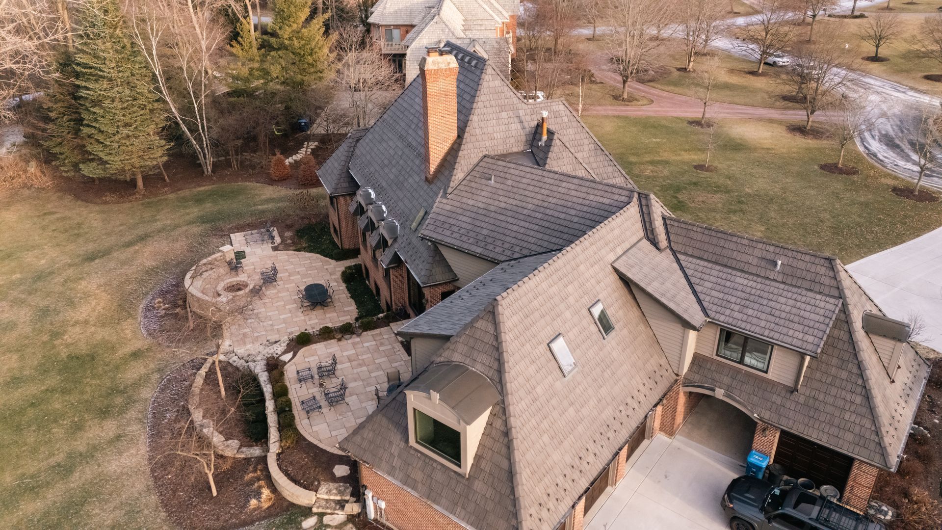 Aerial view of a large, brown roofed house with a brick exterior, a patio, and a black vehicle in the garage.