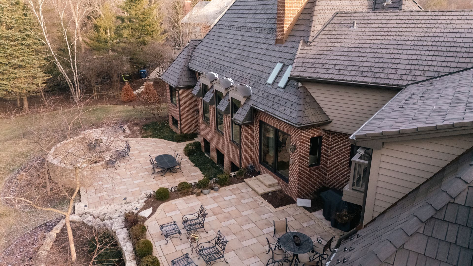 Aerial view of a large brick and wood-sided house with a stone patio, outdoor furniture, and surrounding trees.