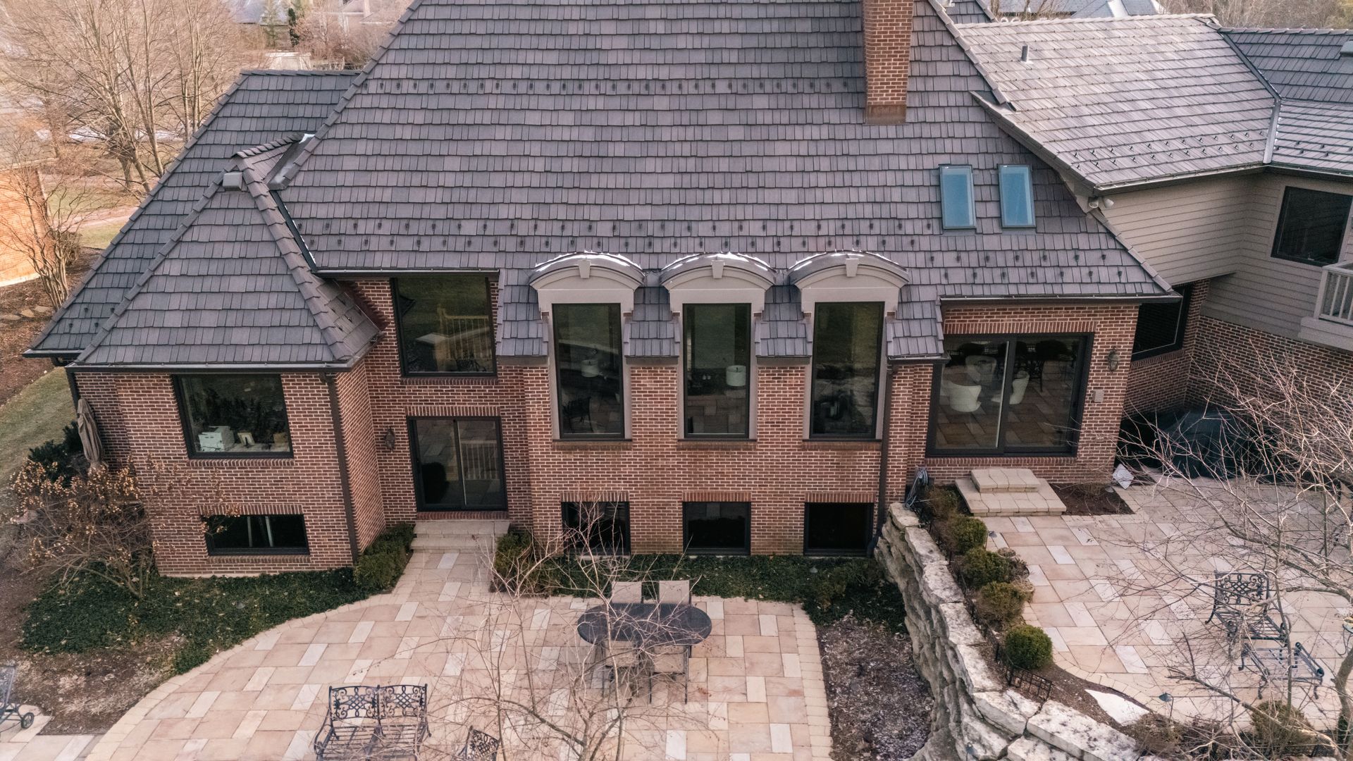 Brick house with gray shingle roof, large windows, patio with table and chairs.