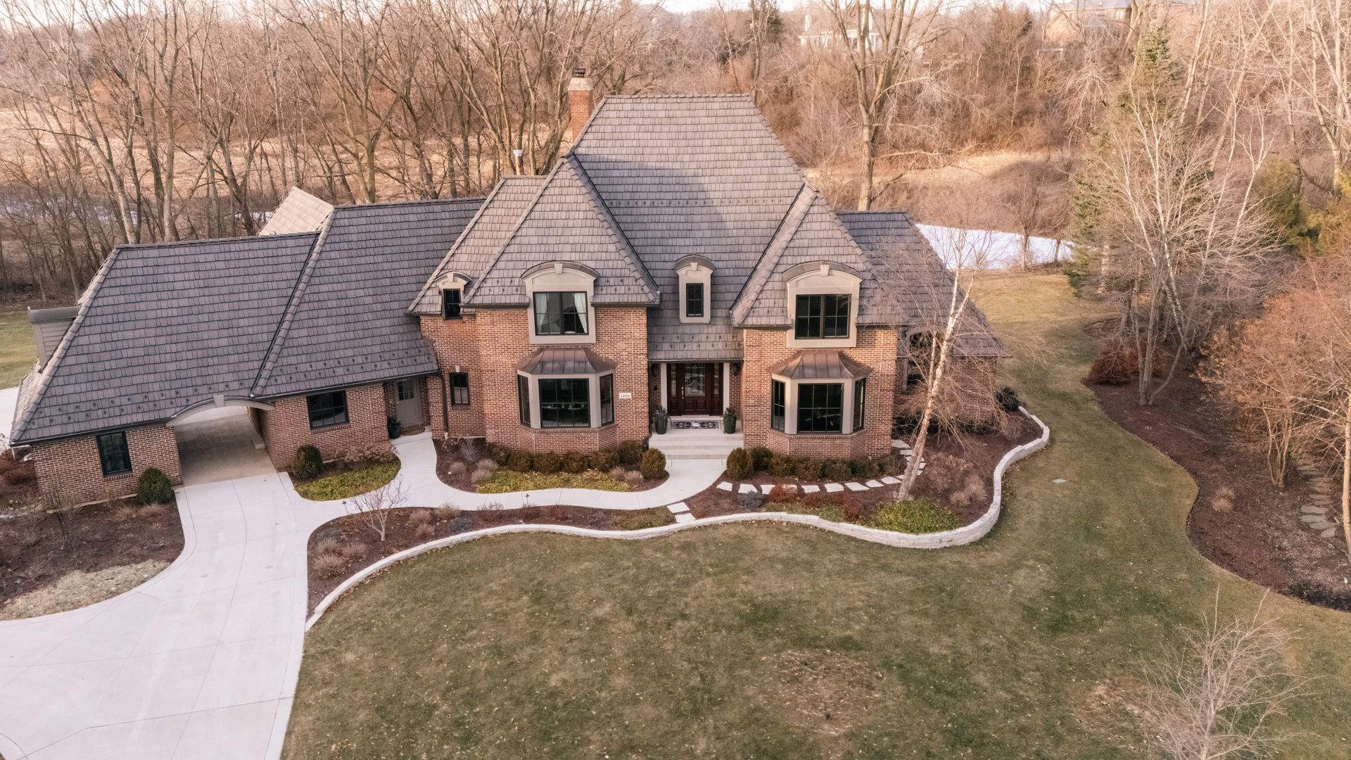 Brick house with gray roof and a long driveway surrounded by trees and grass.
