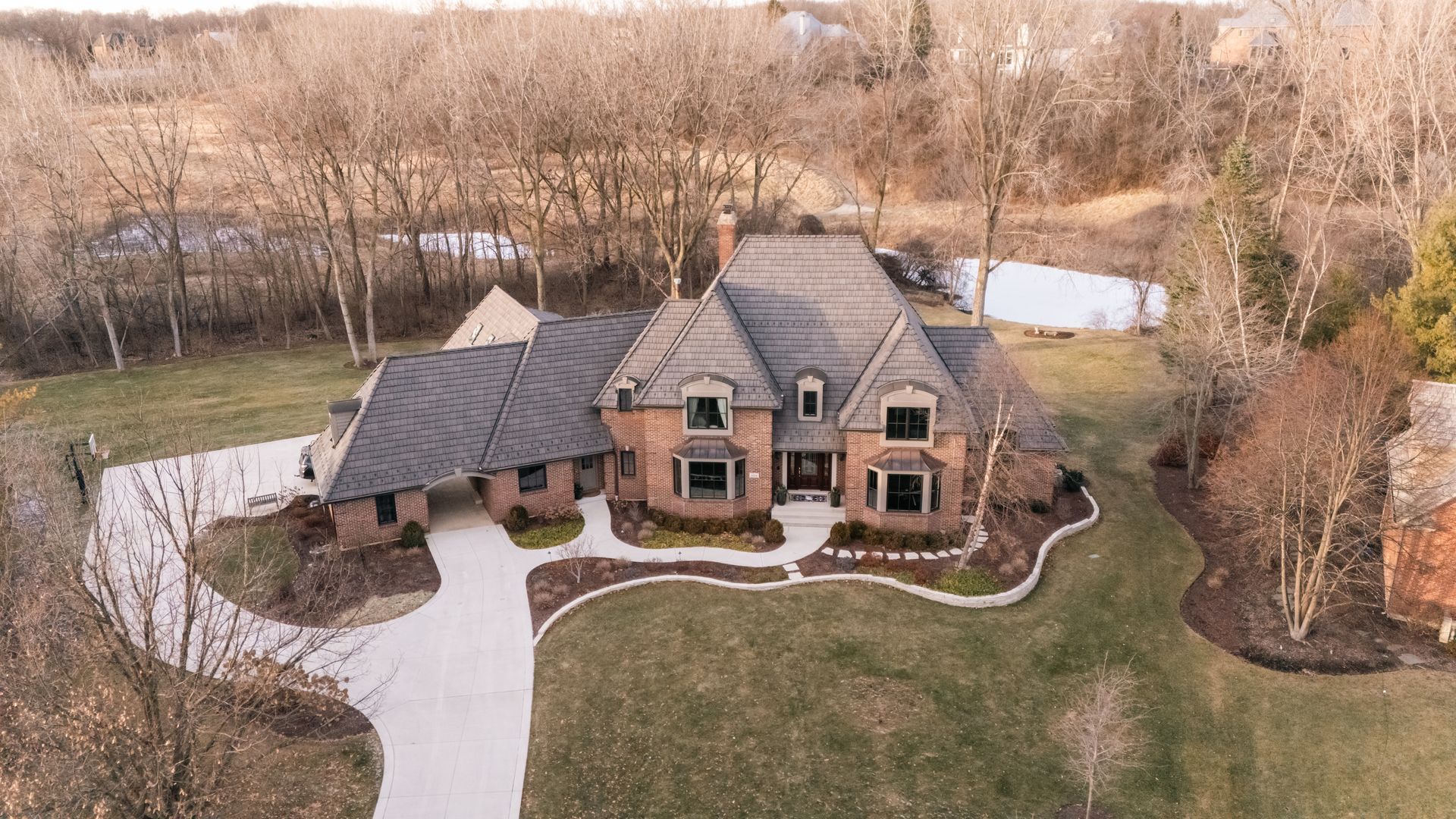 Drone shot of a brick home with a long driveway, surrounded by trees and grass.