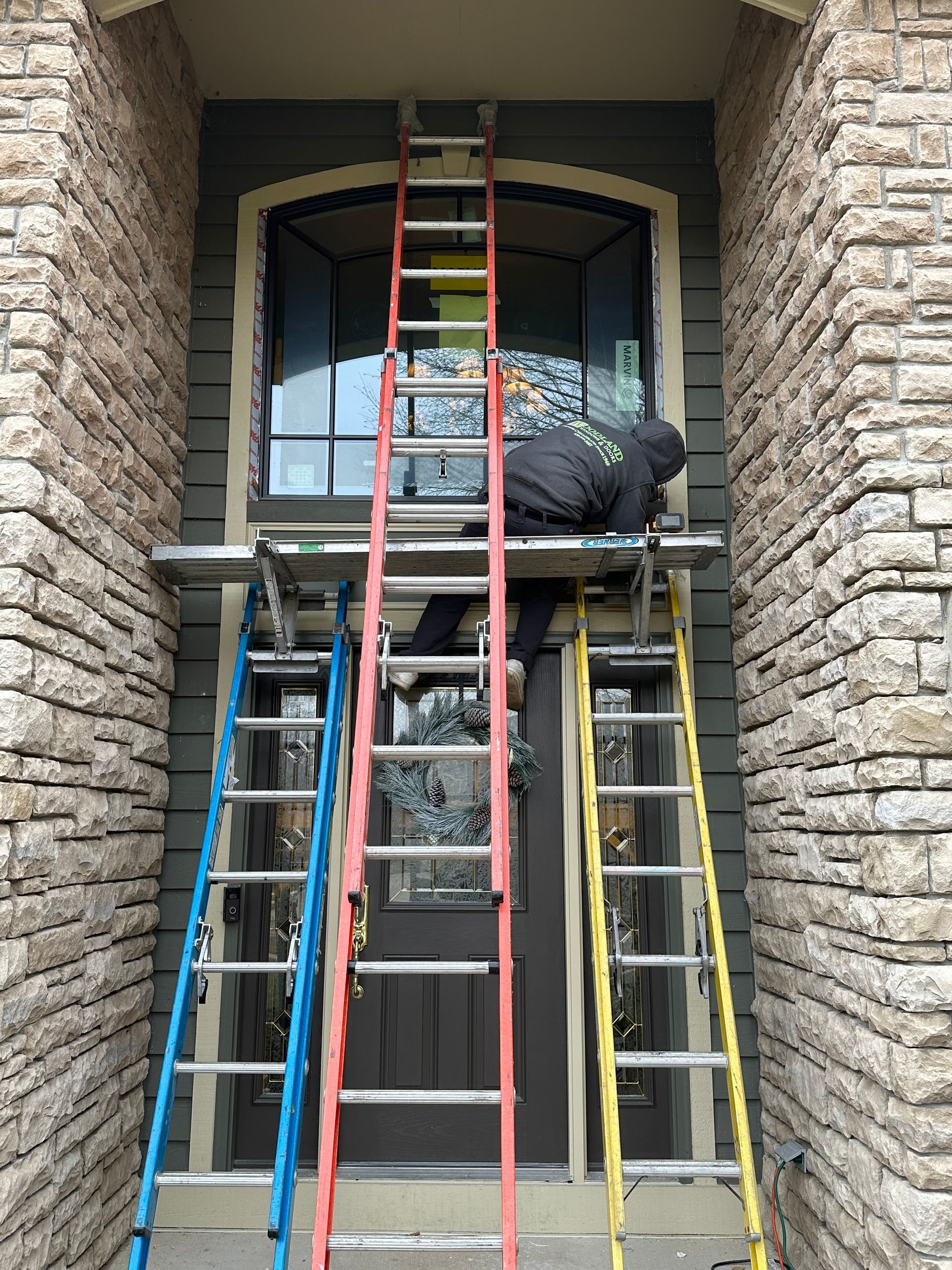 Person working on exterior window, standing on a platform built with three ladders of different colors.