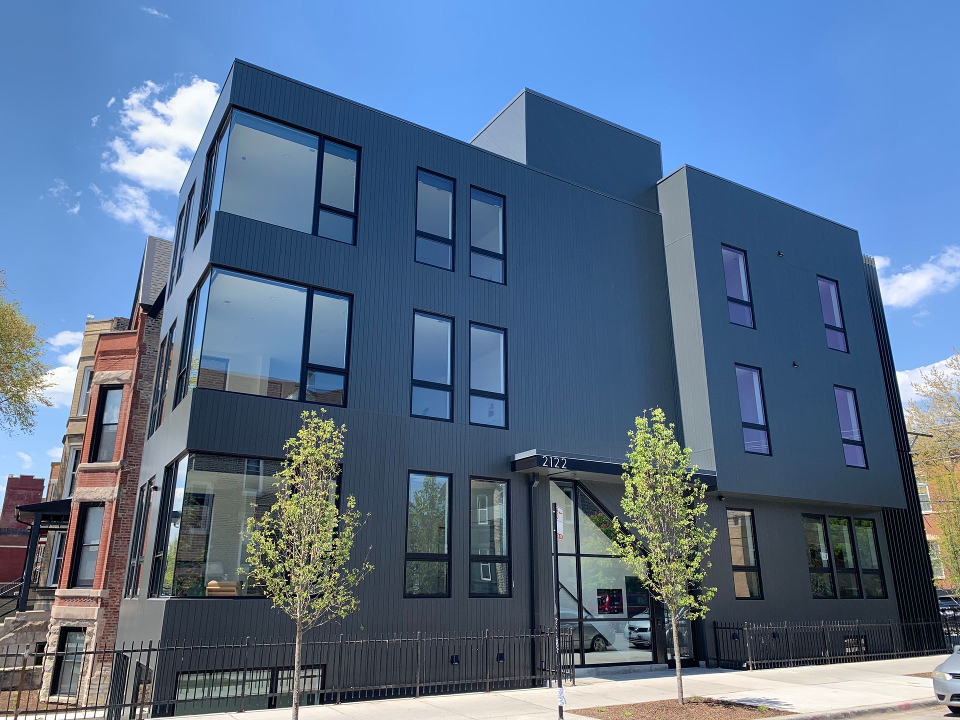 Modern black building with large windows, a small yard and two small trees.